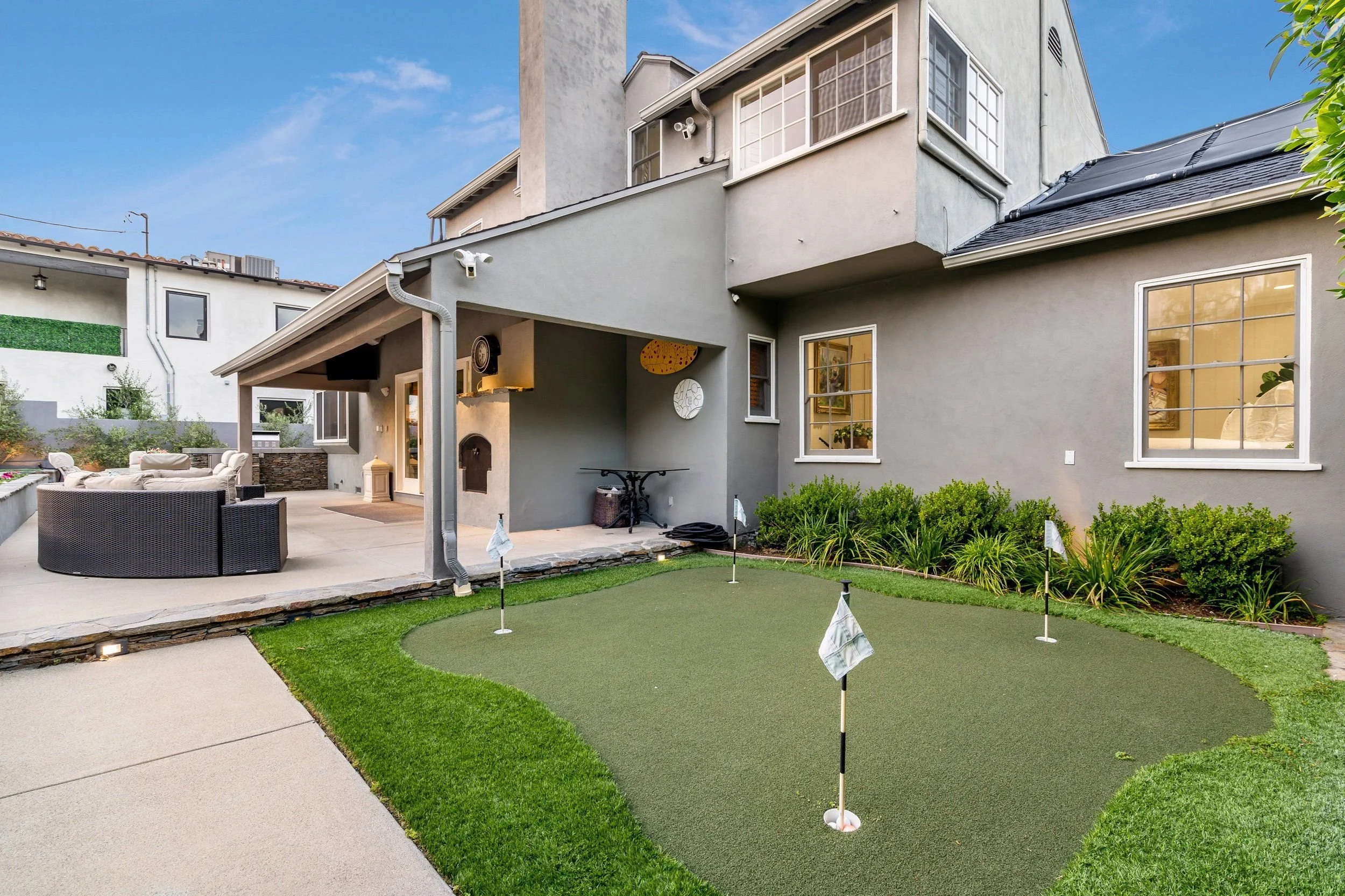 Backyard with a putting green, outdoor furniture, and a two-story house with multiple windows, a small balcony, and exterior surveillance cameras under a blue sky.