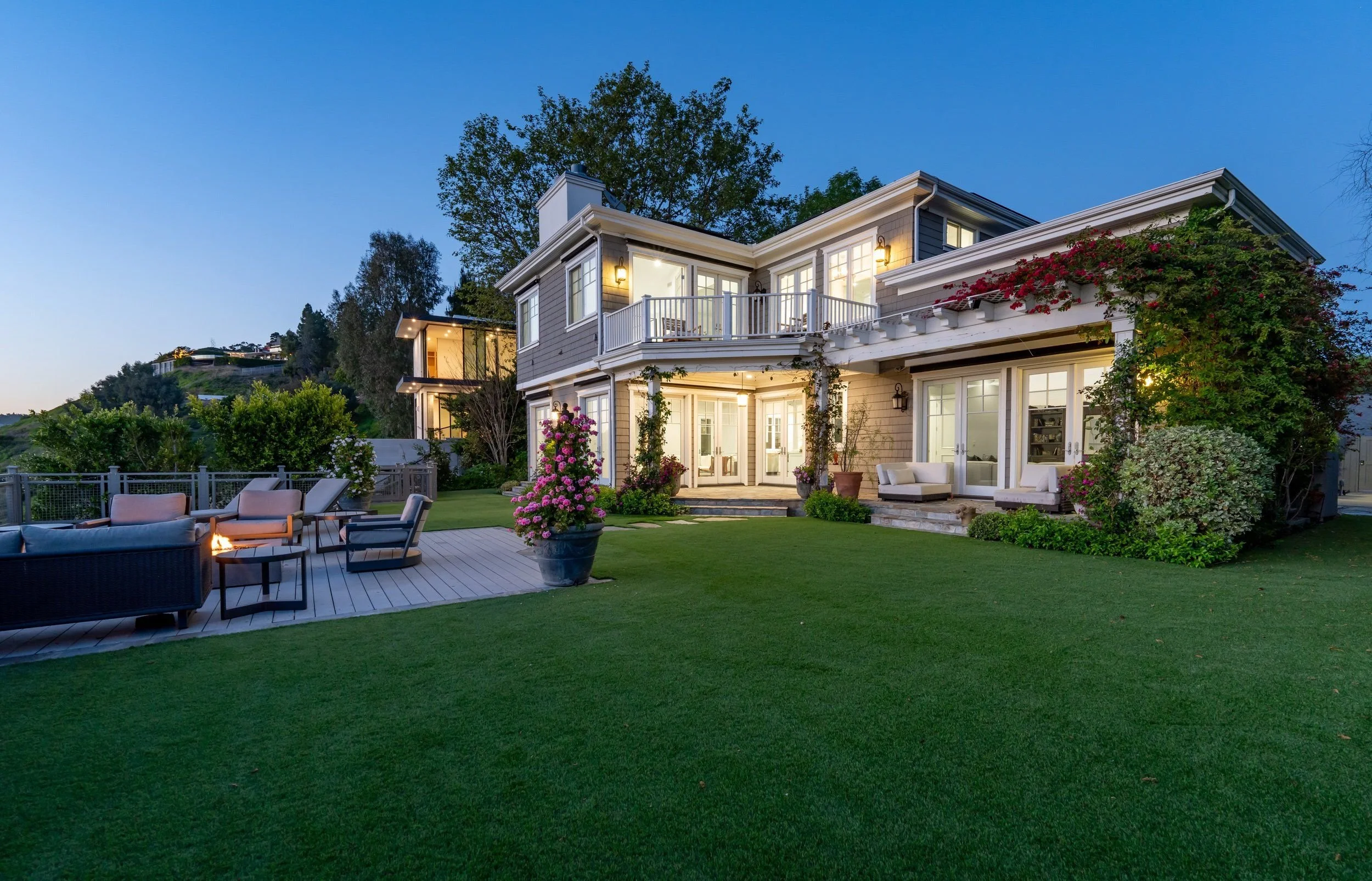 Backyard patio area with outdoor seating, potted plants, and a large two-story house with lit windows, surrounded by greenery and trees at dusk.
