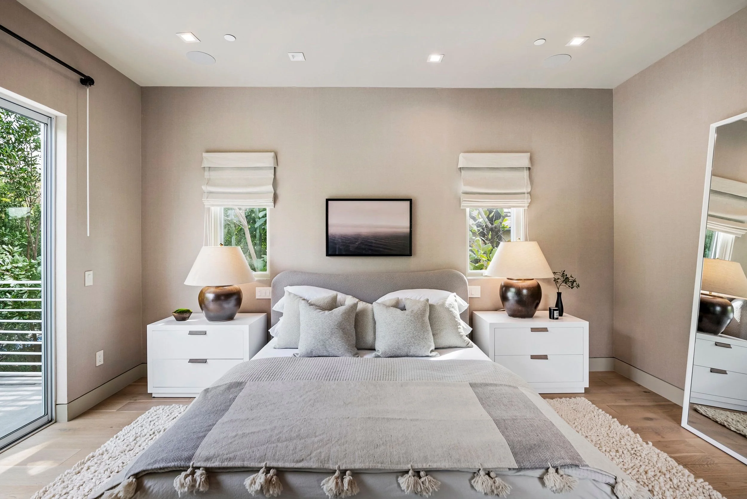 A bedroom with a bed in the foreground, two nightstands with lamps, a wall-mounted TV, and windows with white blinds showing greenery outside.