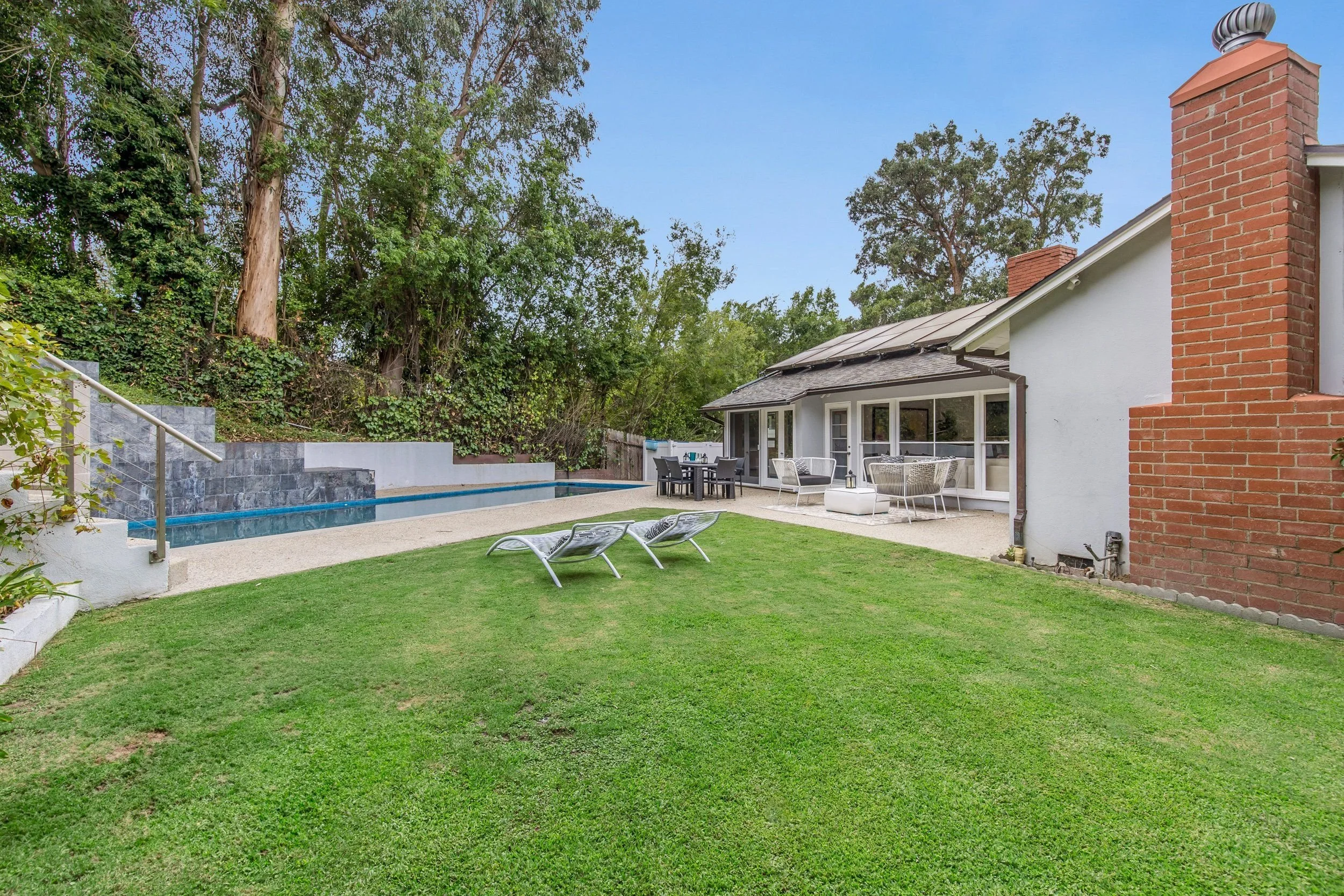 Backyard with a swimming pool, outdoor dining table, lounge chairs, and a house with a brick chimney, surrounded by trees and greenery.