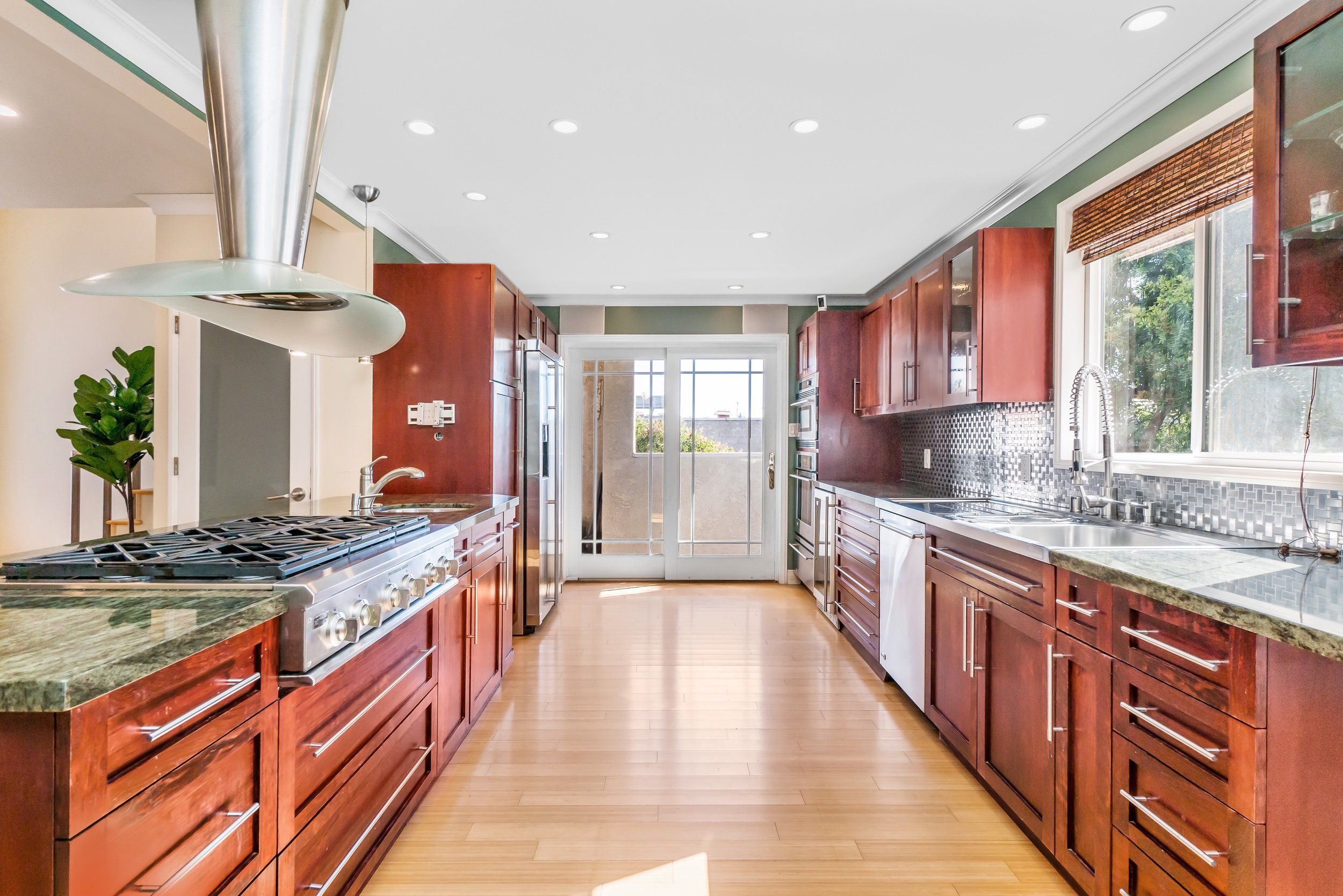 Kitchen with red wooden cabinets, granite countertops, a stainless steel stove with a range hood, a stainless steel refrigerator, and a window with a wooden blind, hardwood floor, and recessed ceiling lights.