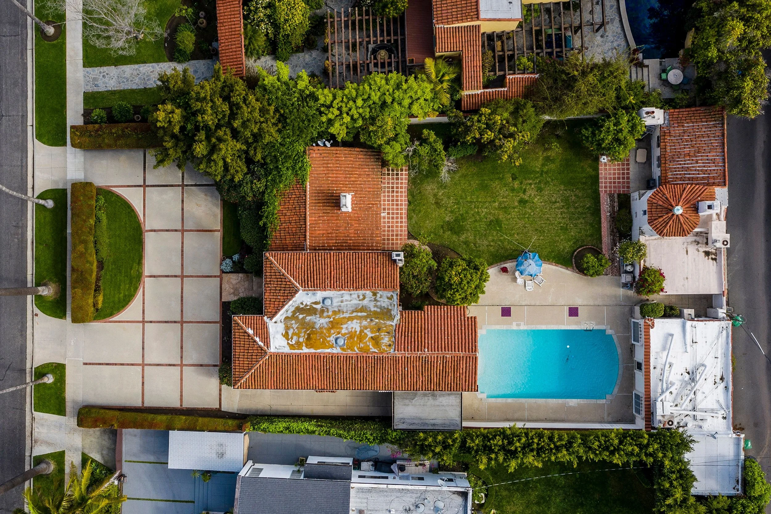 An aerial view of a residential backyard with a large swimming pool, landscaped garden, trees, and various pathways and patios.