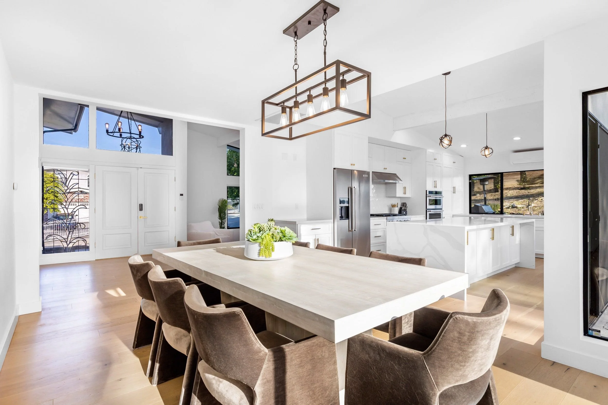 A modern open-concept kitchen and dining area with a large wooden table surrounded by brown upholstered chairs, white cabinets, stainless steel appliances, and contemporary pendant lighting fixtures.