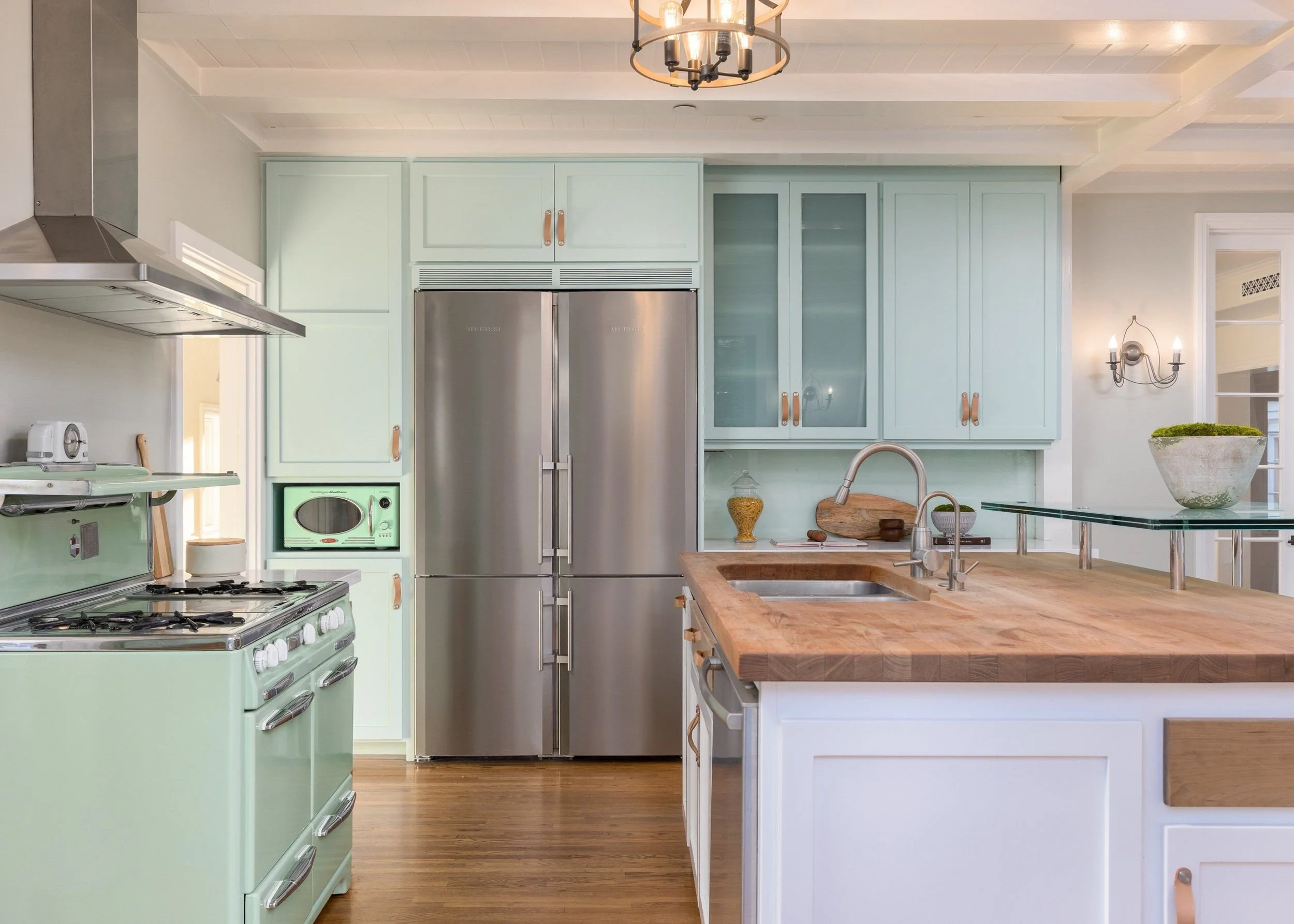 Kitchen with pastel green appliances, wooden countertop island, and light blue cabinets, featuring a stainless steel refrigerator and vintage stove, with a bowl of green moss plant on the side.