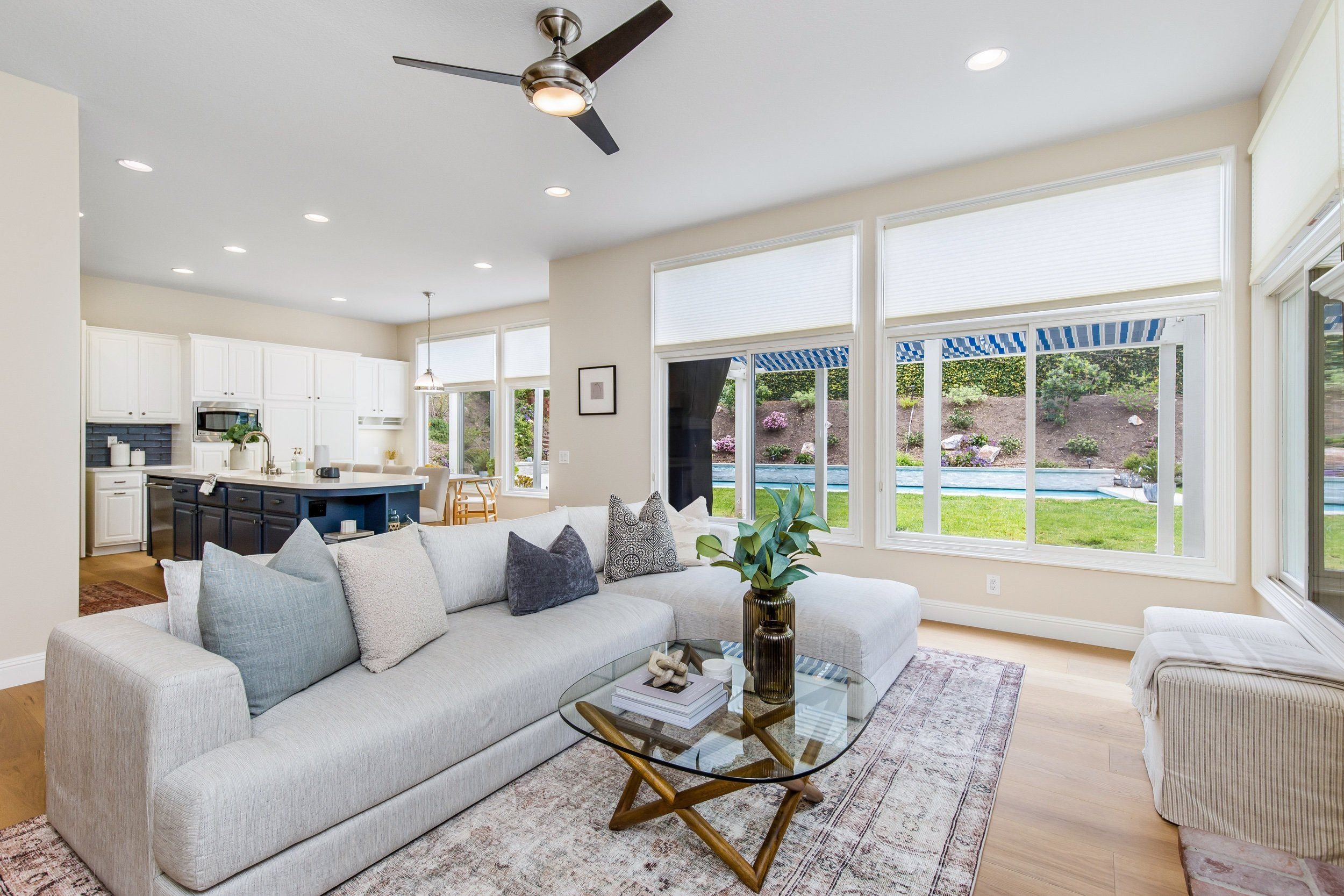 Living room with large windows, white sofa with pillows, a glass coffee table, and view of backyard with pool.