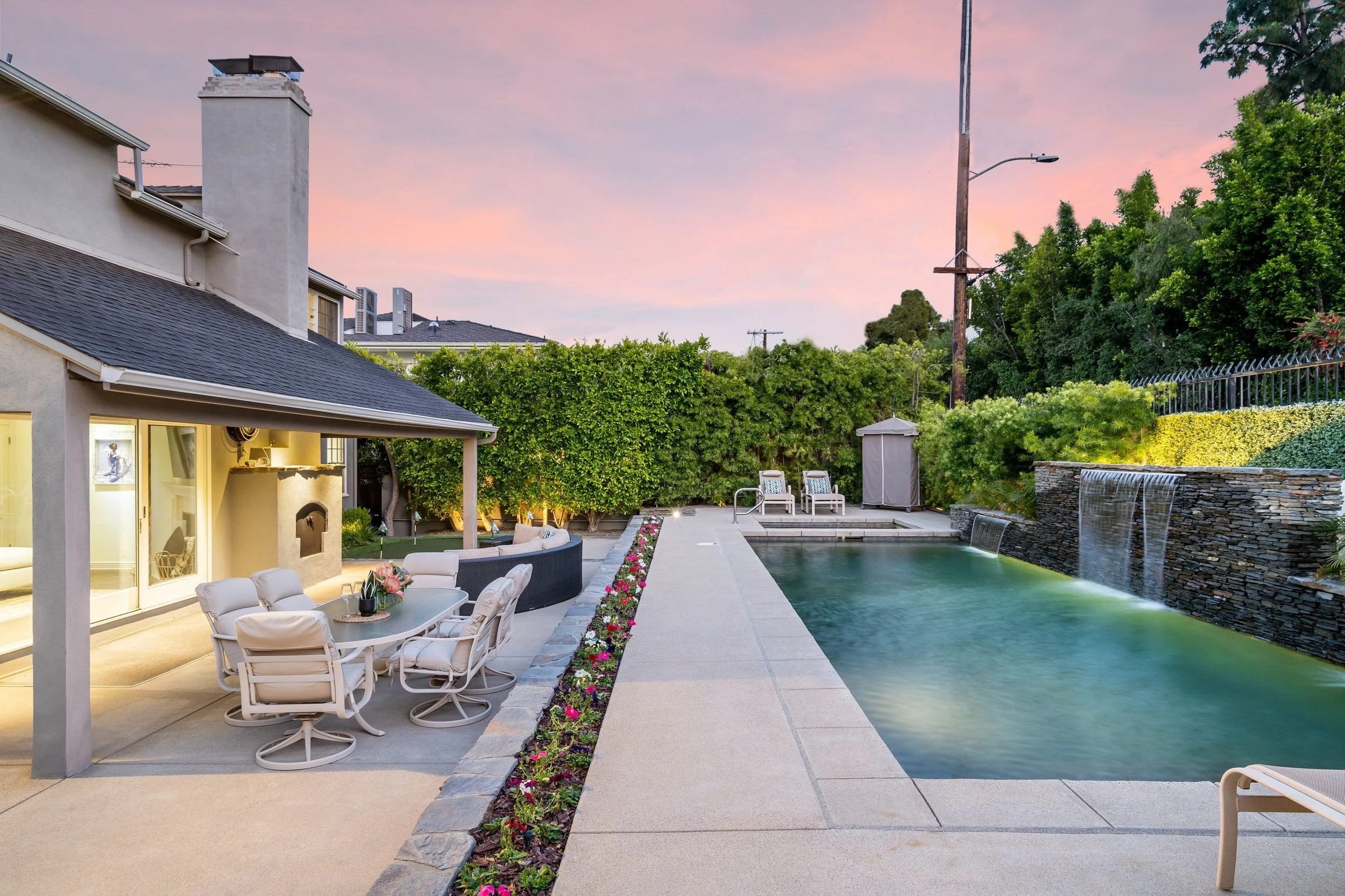 A backyard scene at sunset with a swimming pool, outdoor furniture, lush green trees, and a house with a chimney.