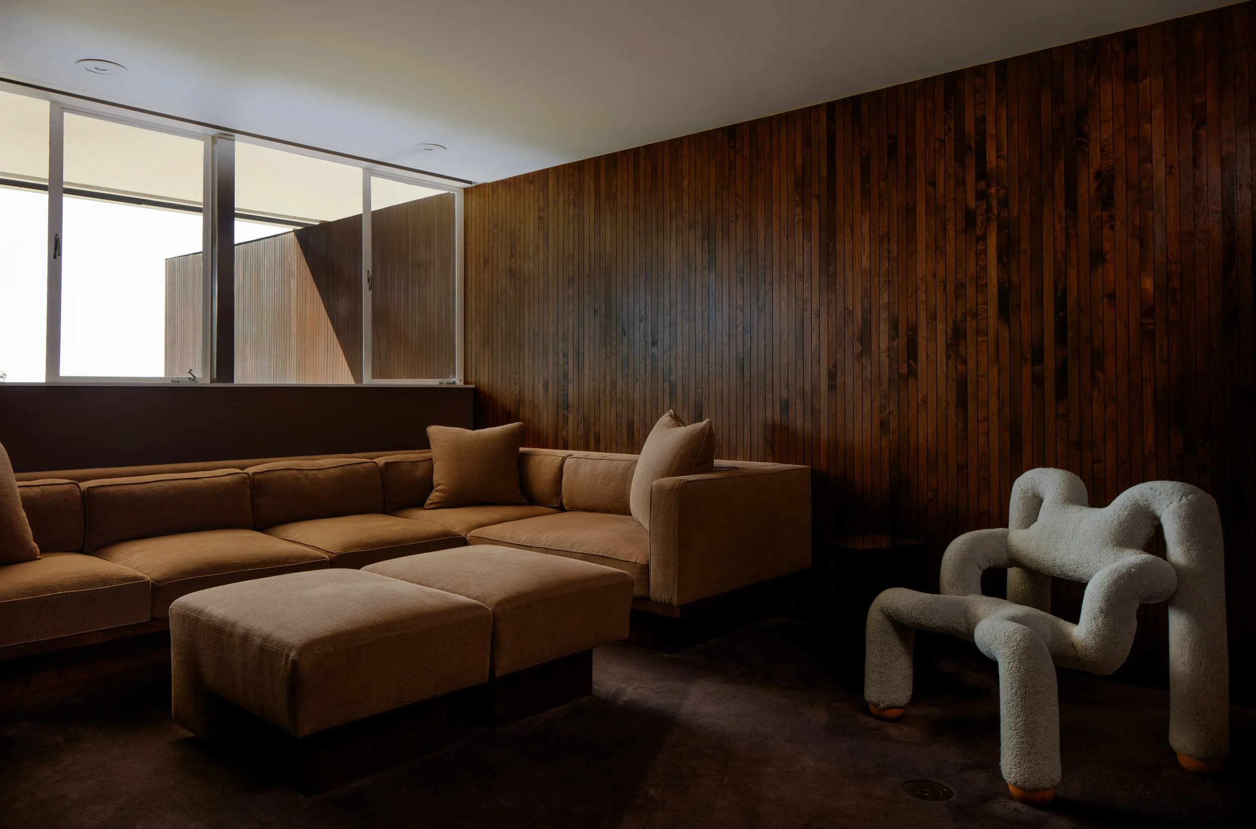 Modern living room with beige sectional sofa, textured plush white armchair, wooden wall panels, large window letting in natural light.