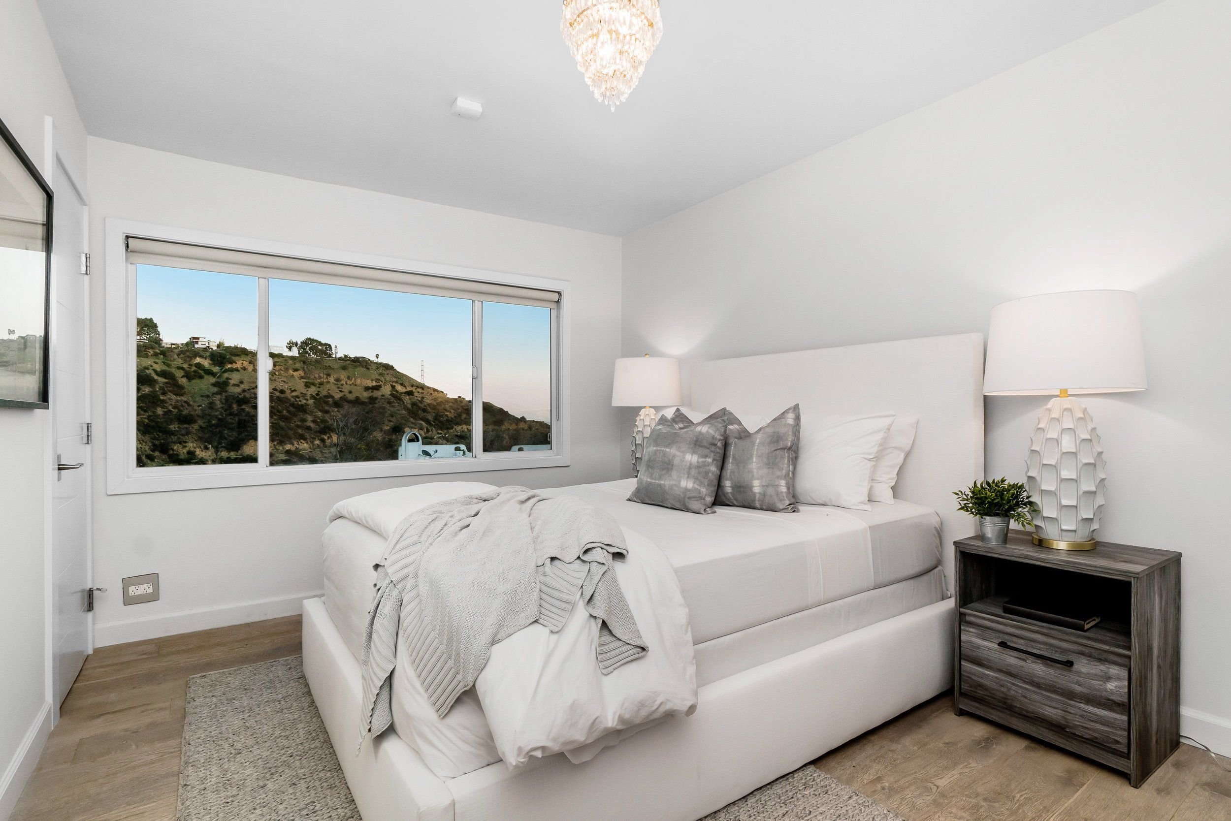 A modern bedroom with white walls, a large bed with white bedding and gray pillows, two matching white lamps on nightstands, a large window showing a hillside landscape, and a chandelier hanging from the ceiling.