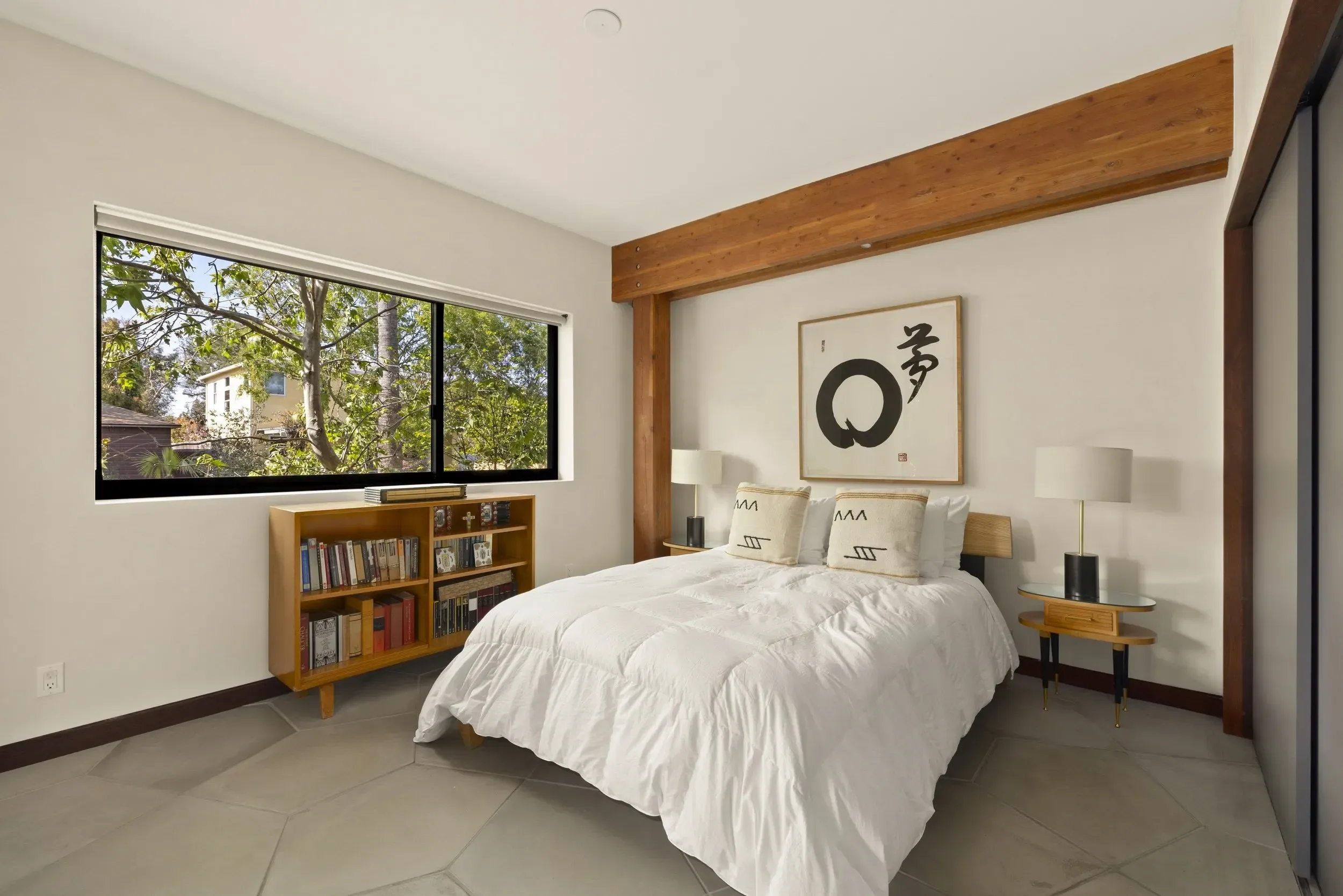 A bedroom with a large window showing trees outside, a wooden headboard with a piece of artwork featuring black calligraphy, two bedside lamps, and a small bookshelf filled with books.