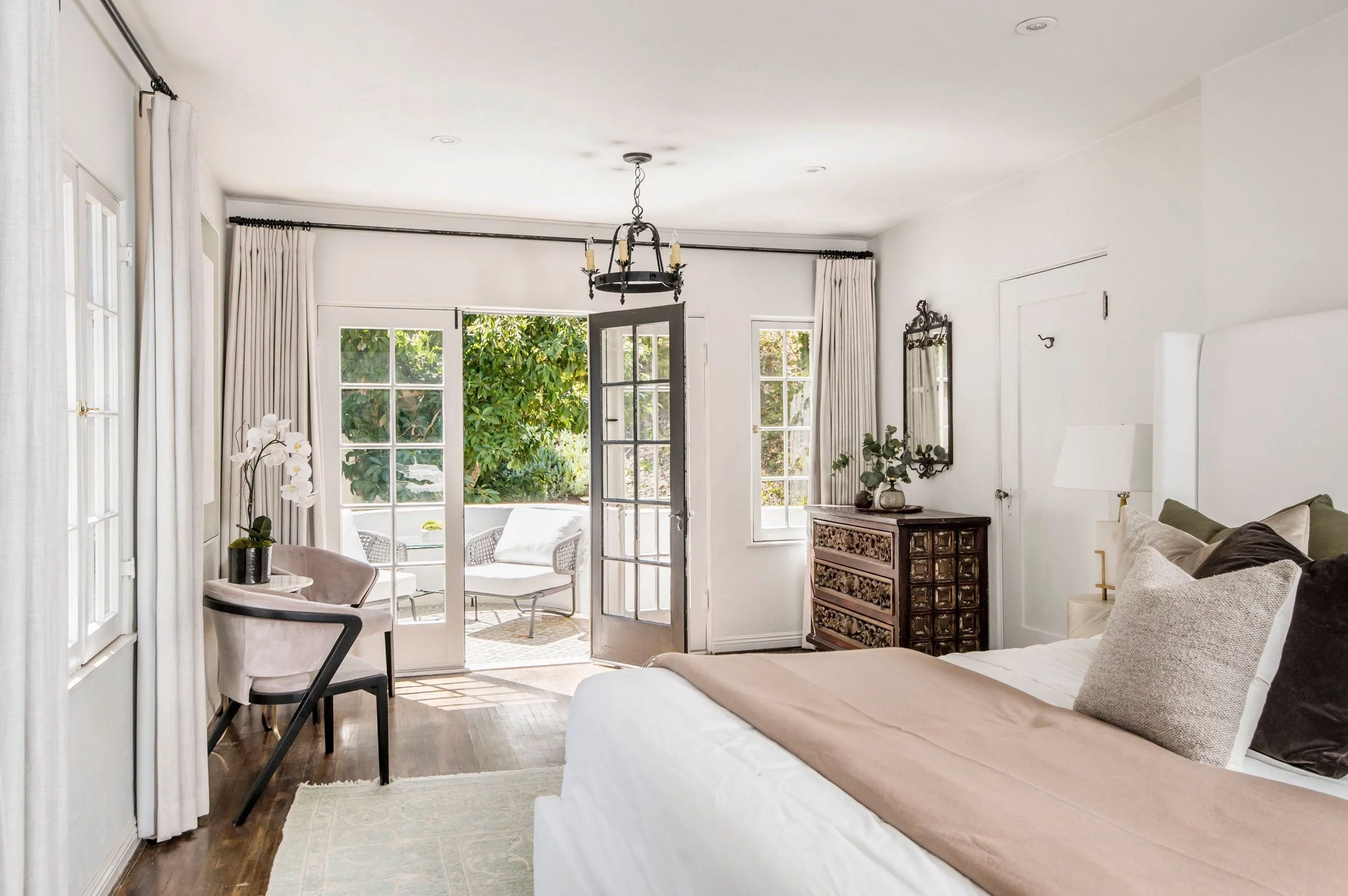 Bright living room with white walls, large windows, and glass doors leading to an outdoor patio. Interior features include a chandelier, a wooden cabinet, a sofa with pillows, a side table with a potted plant, and curtains.