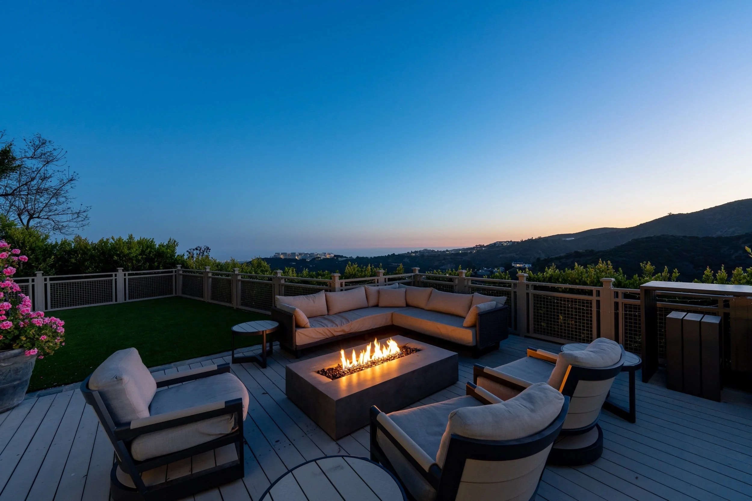 An outdoor deck at dusk with a fire pit in the center, surrounded by white cushioned chairs and a sectional sofa, overlooking rolling hills and a distant cityscape with a clear blue sky.