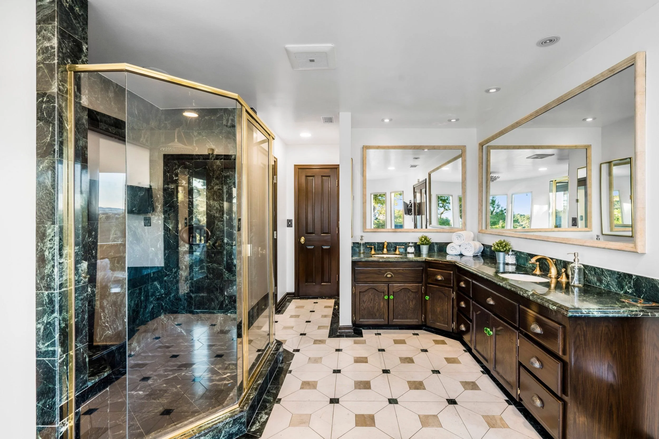 A bathroom with a glass shower enclosure, a wooden vanity with a green marble countertop, and multiple mirrors with wooden frames. The room has a black and white tiled floor, and windows showing a view of trees outside.
