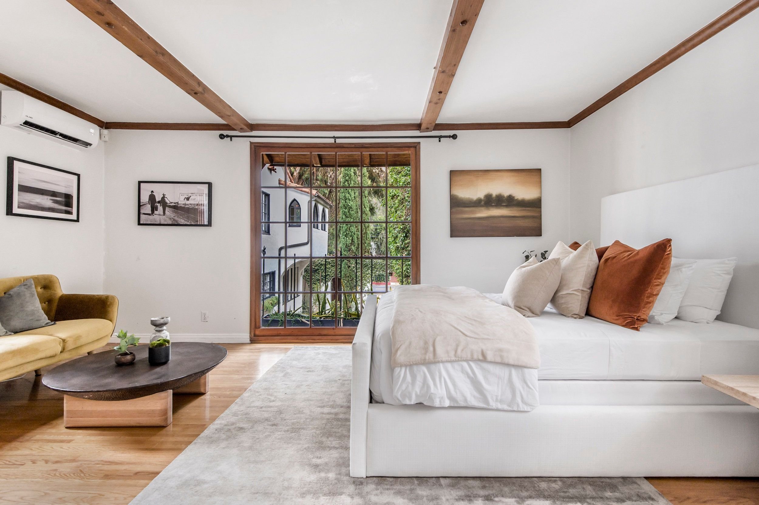 Living room with white walls, wooden beams on ceiling, large window, yellow and white couches, framed photographs, and artwork.