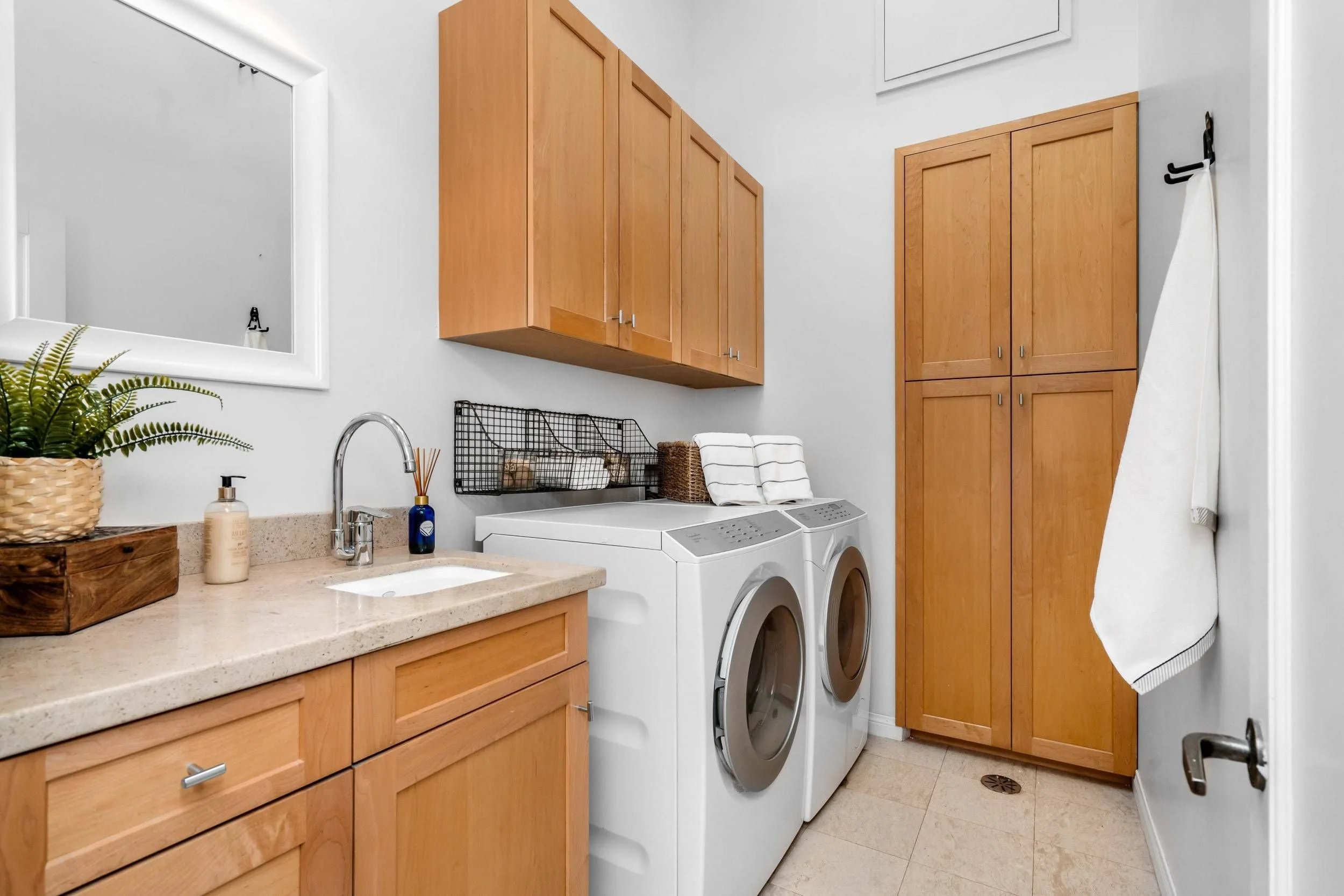 Laundry room with wooden cabinets, a sink, a potted plant, laundry detergents, a washer and dryer, and a wall-mounted wire basket for laundry essentials.