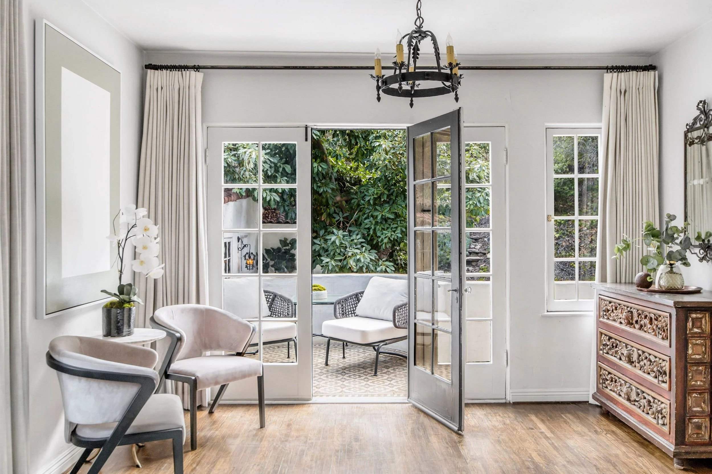 Living room with white walls, a chandelier, a large window, and a glass door leading to an outdoor area with greenery and seating.