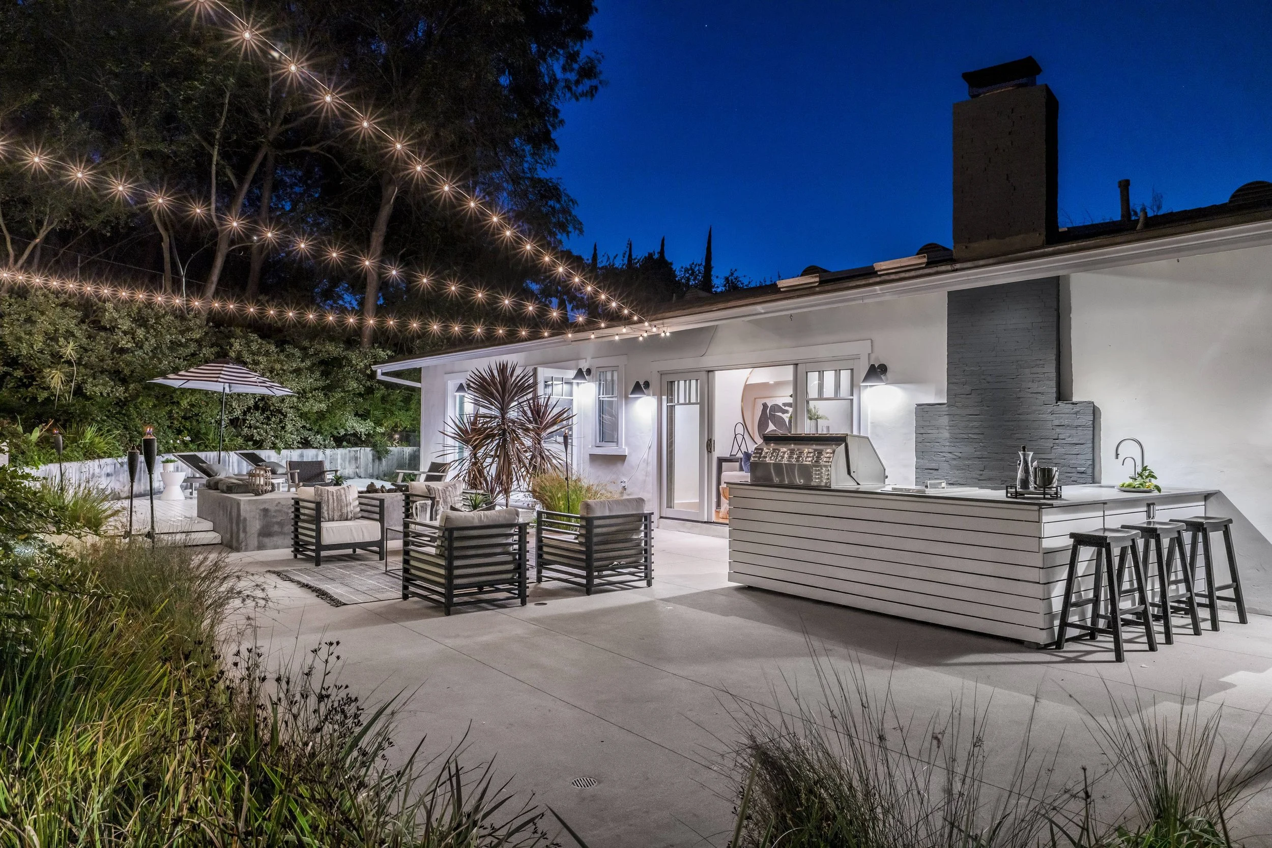 Backyard patio at night with string lights hanging above. Outdoor furniture, including a dining table, chairs, an umbrella, and a kitchen counter with stools. Trees and greenery surround the area. The house has large windows and a chimney, with inter