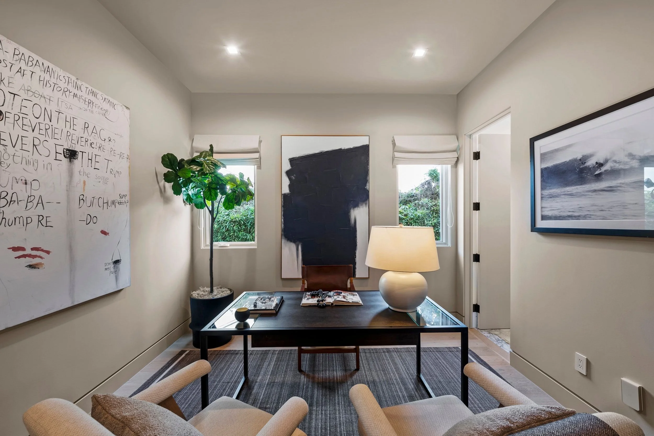 Interior of a modern living room with white walls, two windows with shades, framed artwork, a large potted plant, a table with books and decorative items, a white vase, and a comfortable beige armchair.