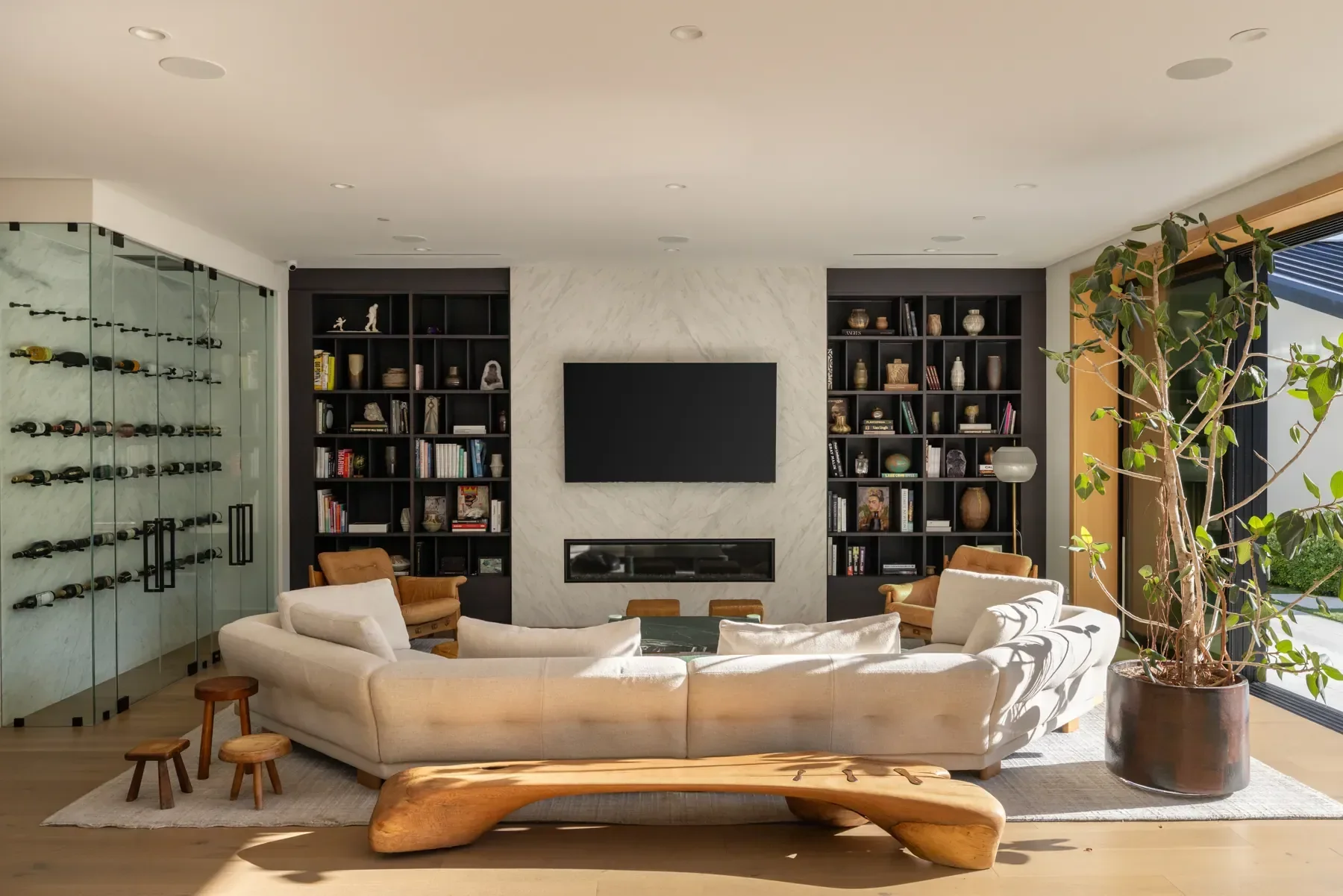 Living room with a curved cream sofa, wooden stools, a large black bookshelf, a wall-mounted TV, and a large potted plant near a window.