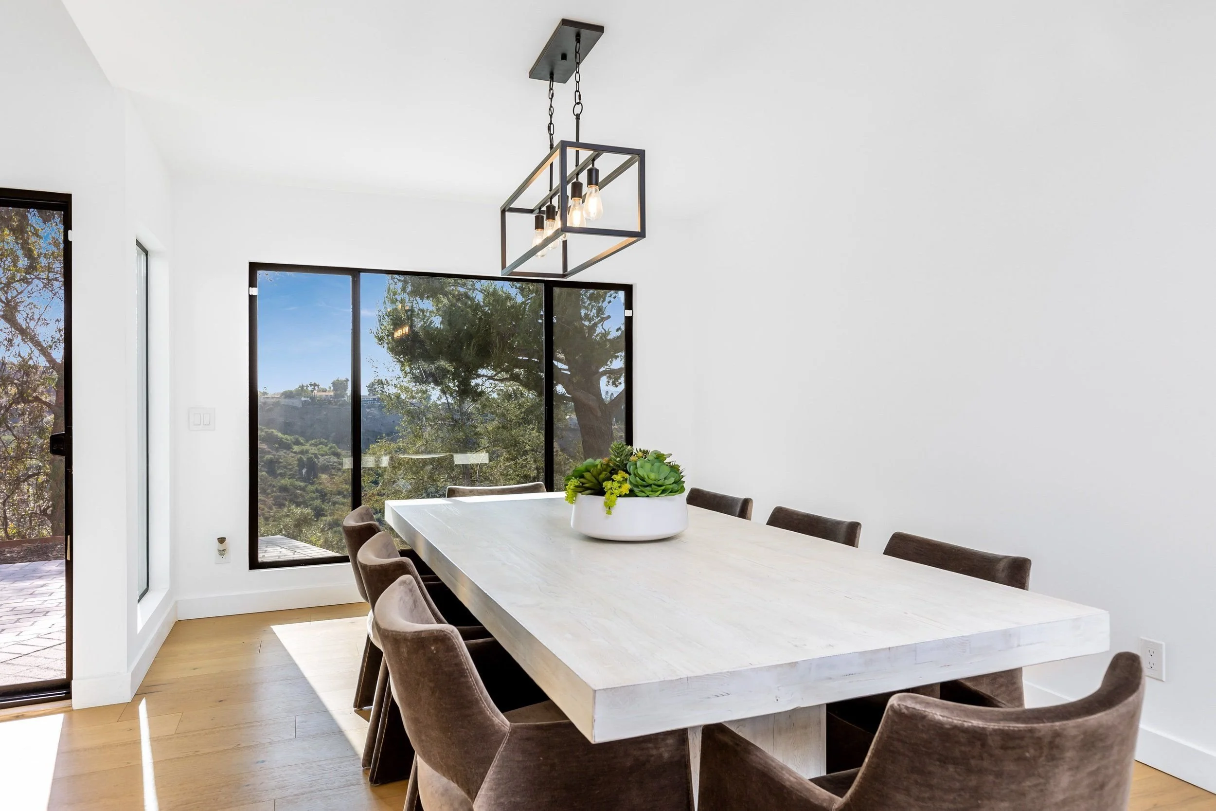 Modern dining room with a rectangular table, upholstered chairs, a decorative plant centerpiece, large windows, and a contemporary pendant light fixture.