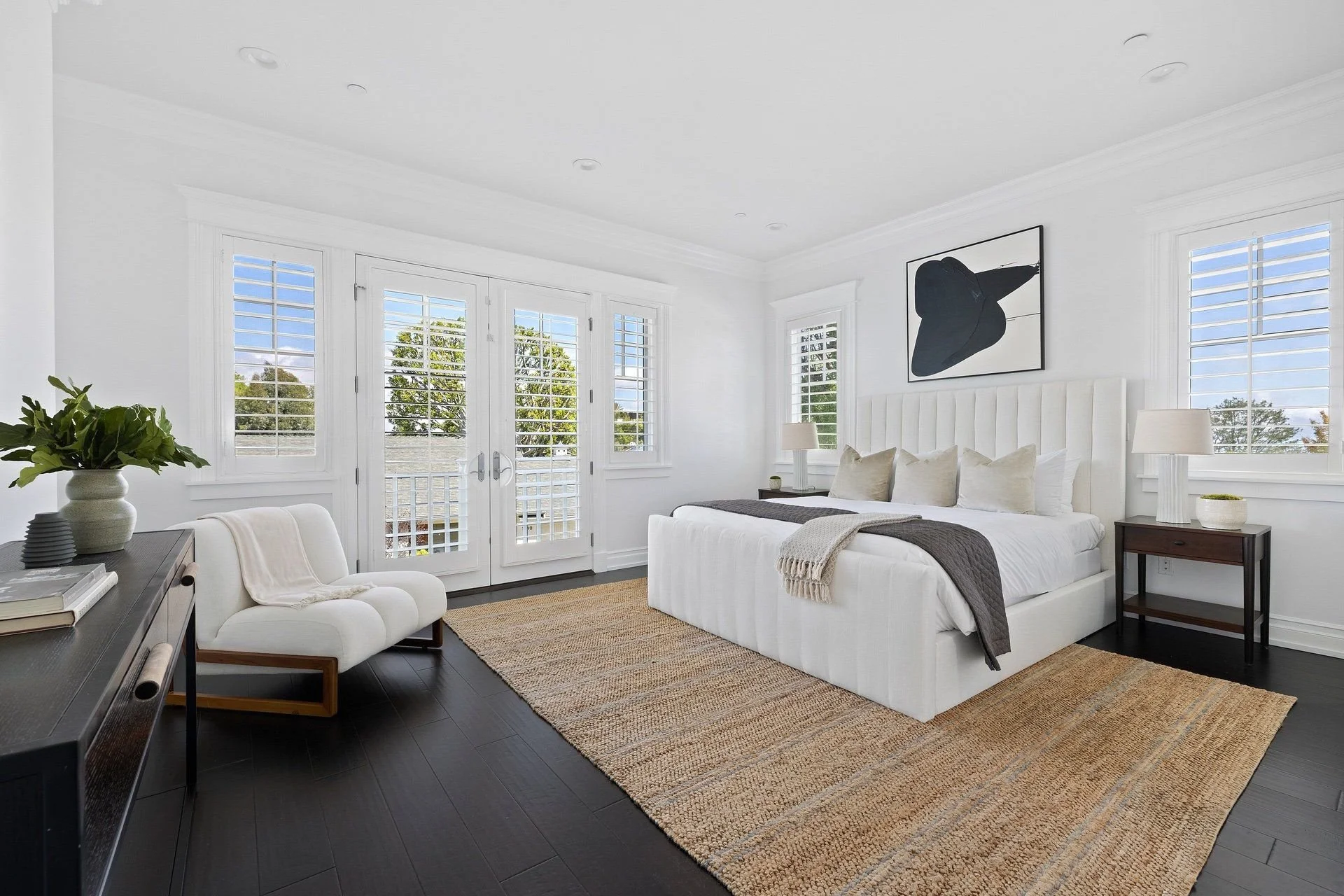Bright bedroom with white walls and ceiling, large windows with shutters, a white bed with beige and gray pillows, nightstands with lamps, a framed black and white hat illustration, a beige rug, a side table with plants, and a white armchair.