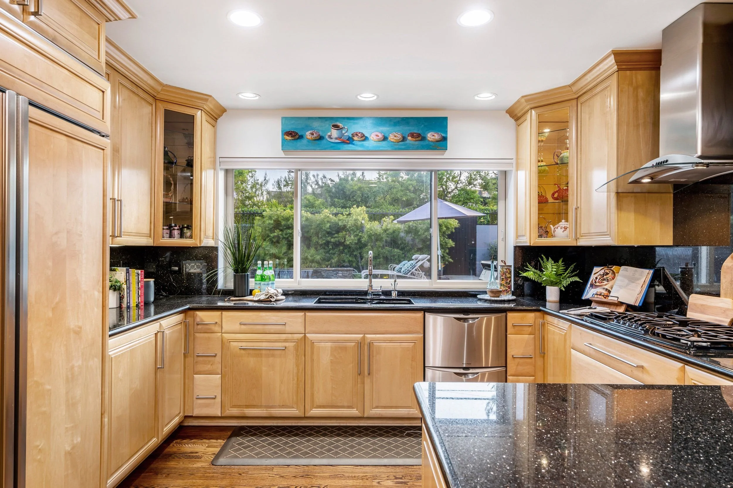 A modern kitchen with light wood cabinets, black granite countertops, and a large window overlooking a backyard with a patio umbrella. The kitchen wall has a painting of donuts and a cup of coffee.