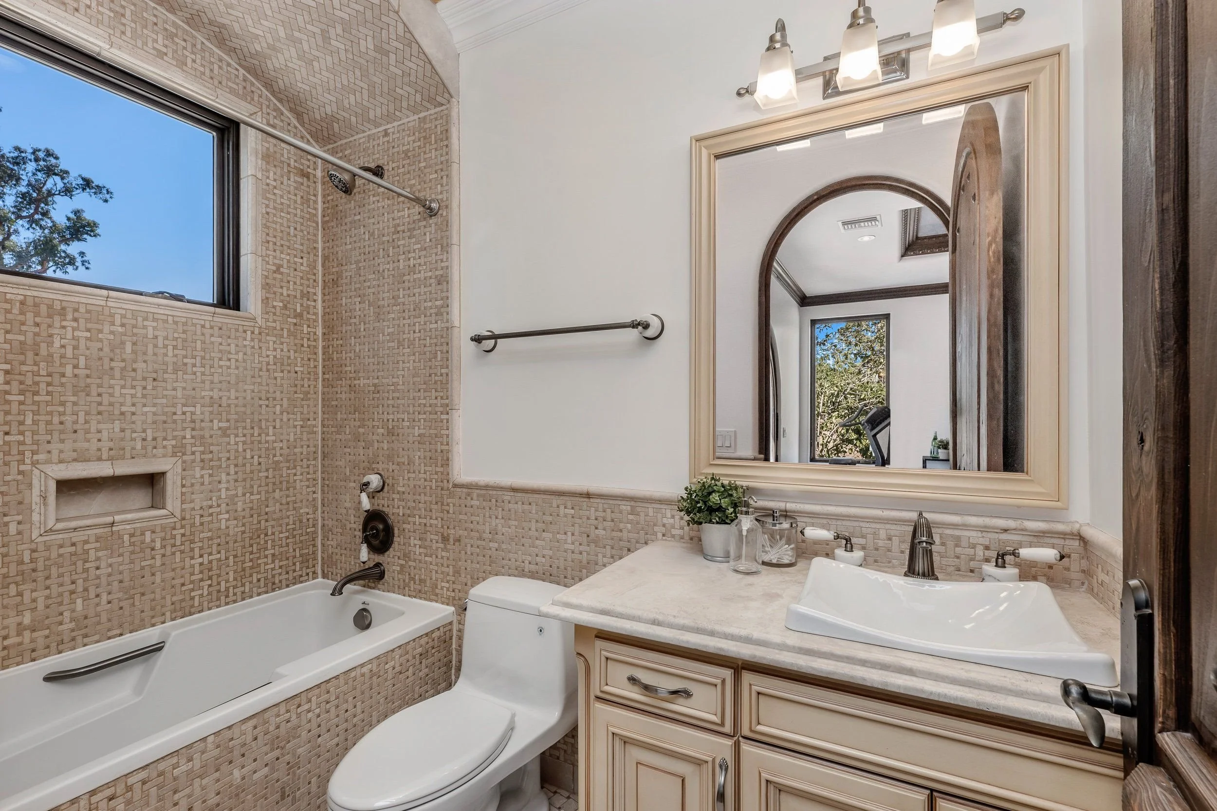 Bathroom with a bathtub, a toilet, a vanity with a sink, a large mirror, and a window showing trees outside. The bathroom features beige textured tile walls and dark wooden accents.