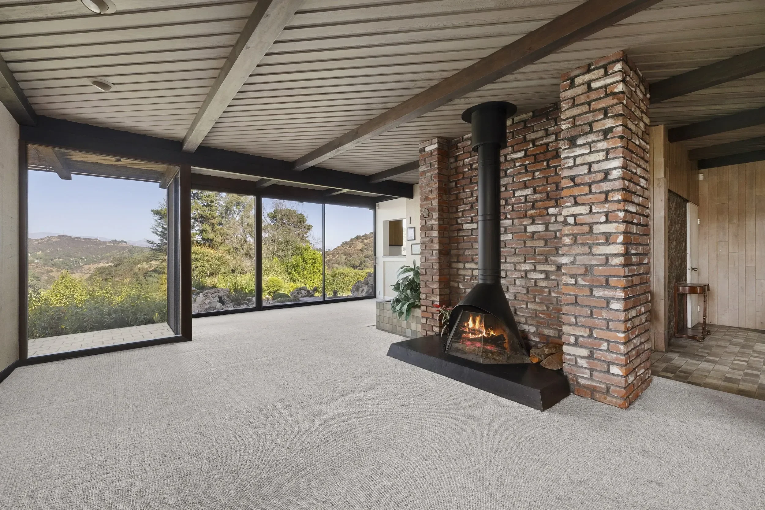 The interior of a modern house with large windows showing a scenic landscape, a brick fireplace with a chimney, and beige carpet flooring.