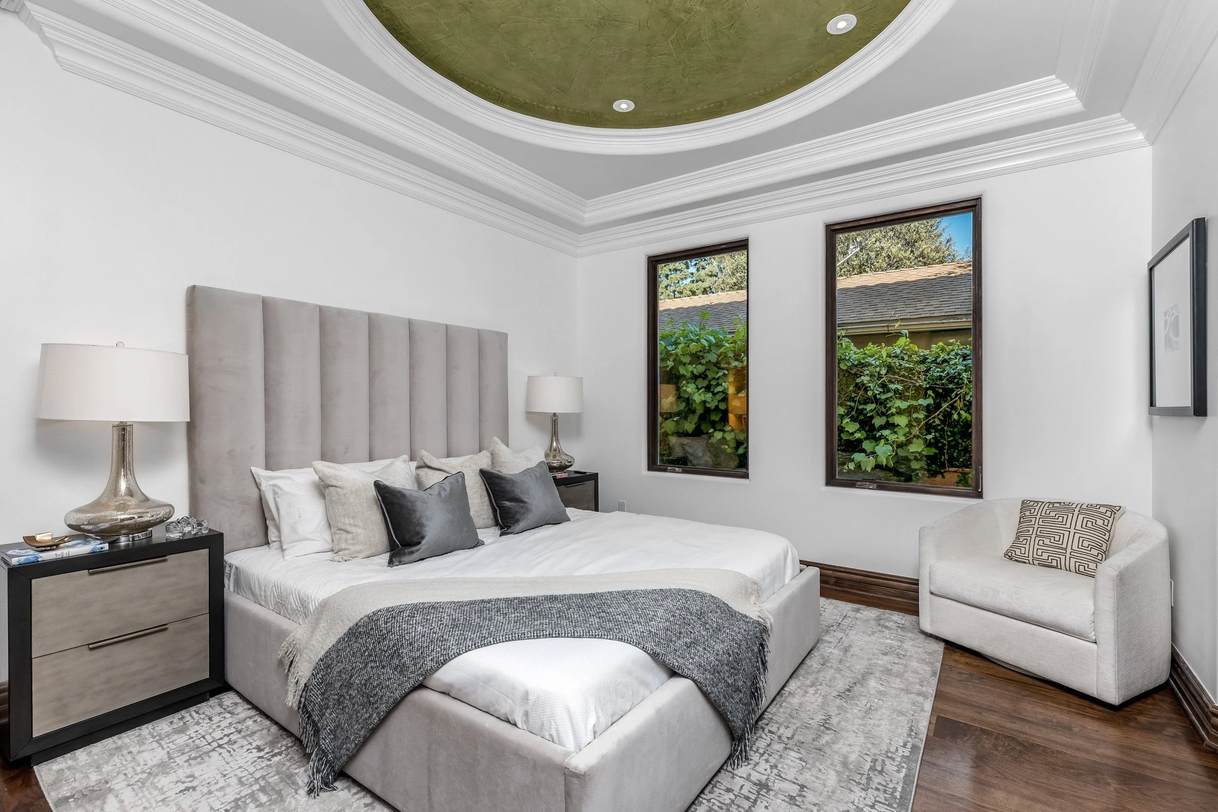 Elegant bedroom with white walls, a large upholstered bed with gray and white pillows, a black nightstand with a lamp, windows with view of greenery, and ceiling with decorative crown molding and a central painted area.