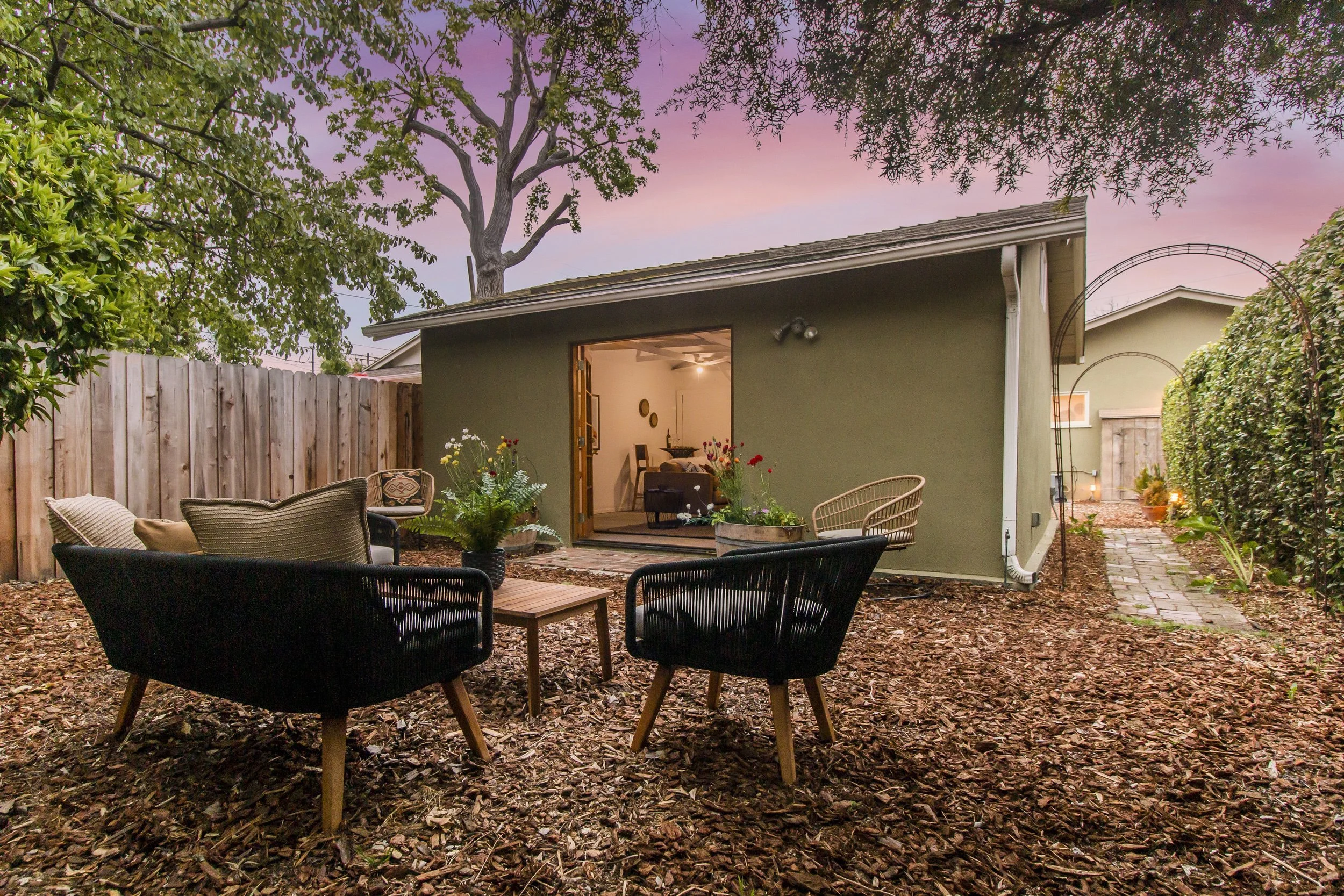 A backyard patio with outdoor seating, wooden fence, and a small shed, surrounded by trees and plants, at sunset with a pink and purple sky.