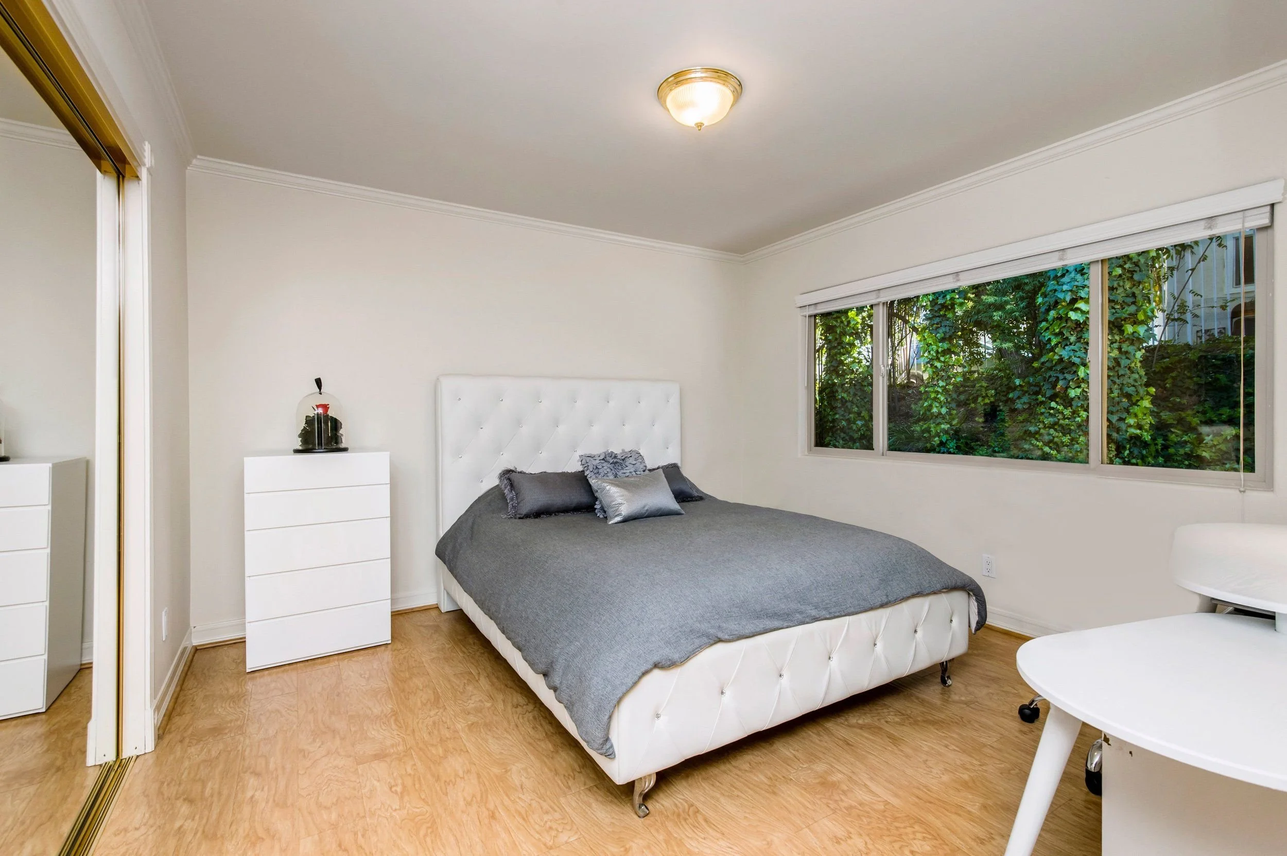 A bedroom with a large window showing greenery outside, a bed with gray bedding and pillows, two white dressers, and a ceiling light fixture.
