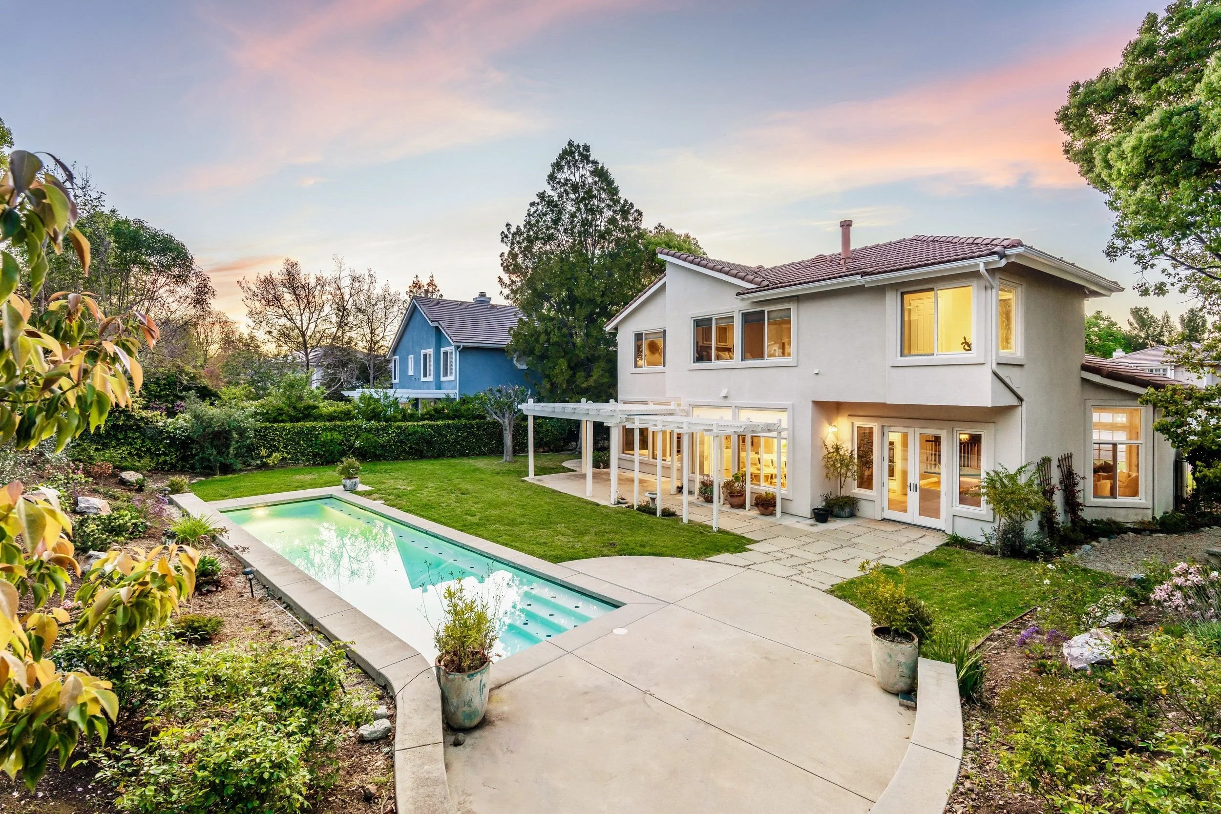 A backyard with a swimming pool, green lawn, and potted plants. A two-story house with large windows and a tiled roof is visible, with indoor lighting on. The sky shows a sunset with pink, purple, and blue hues, and there are trees surrounding the ya