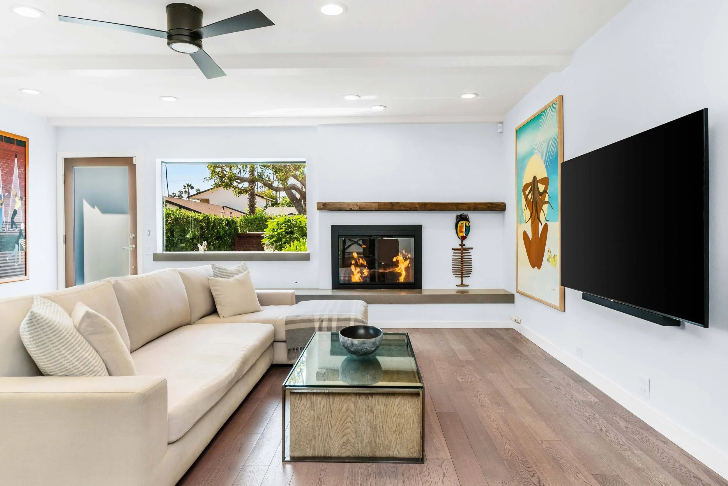 Modern living room with a white sectional sofa, glass coffee table, fireplace, artwork, large window with greenery outside, ceiling fan, and flat screen TV.