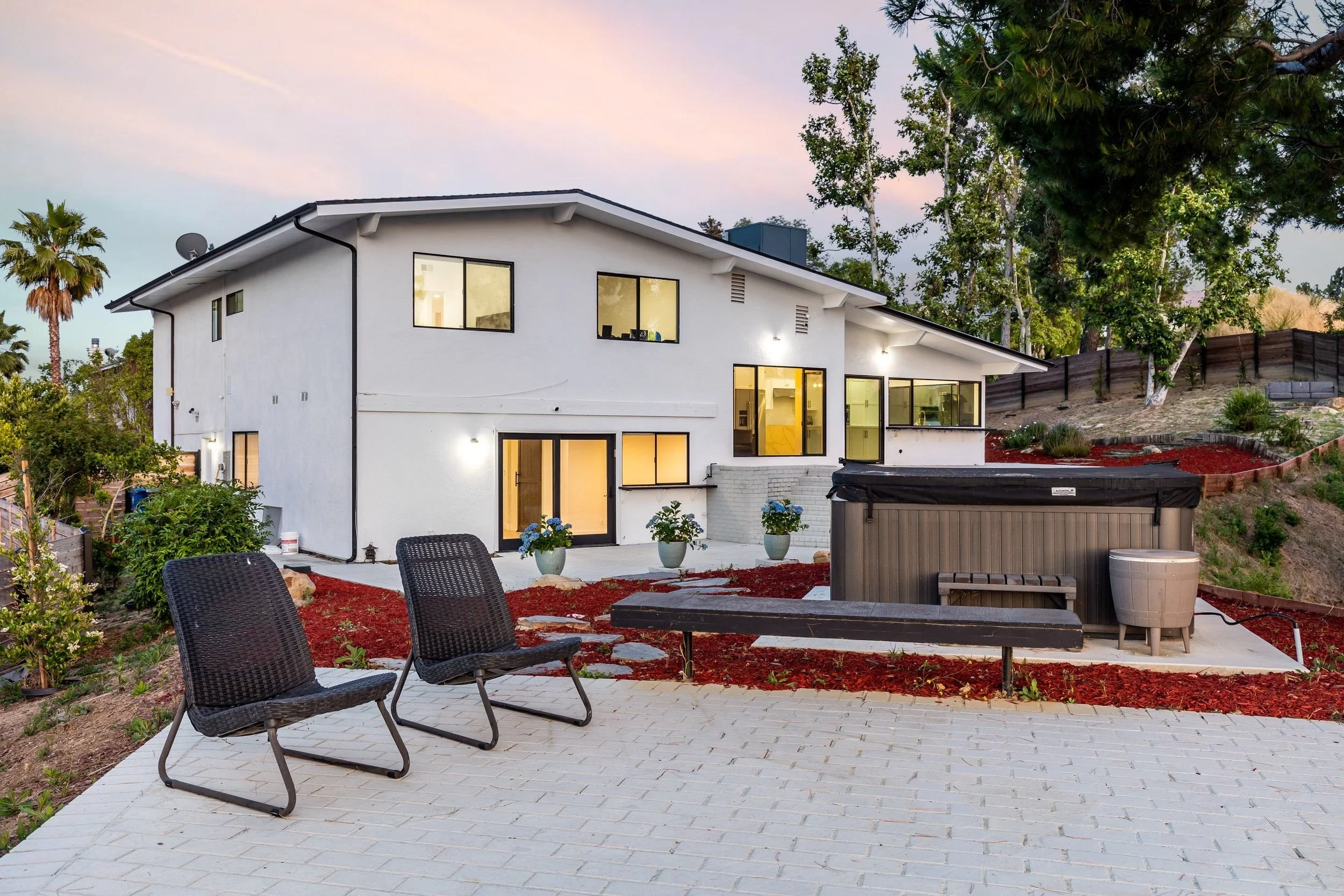 A modern two-story white house with large windows, surrounded by a landscaped backyard with sitting area, chairs, and gardening beds, at dusk.