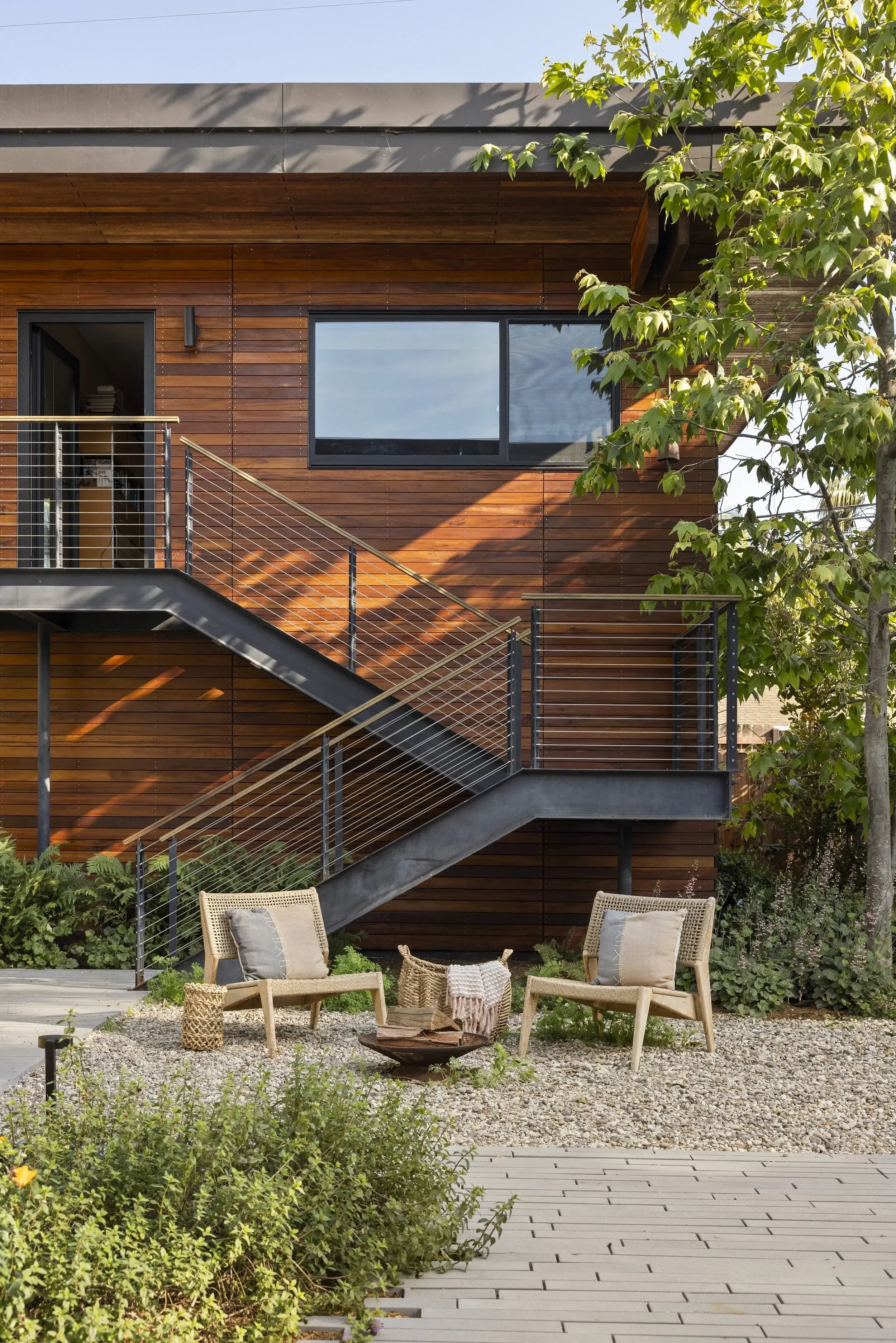 Modern wooden house with large window and an outdoor staircase. There are patio chairs and a small table with a plant in the yard, surrounded by greenery and gravel.