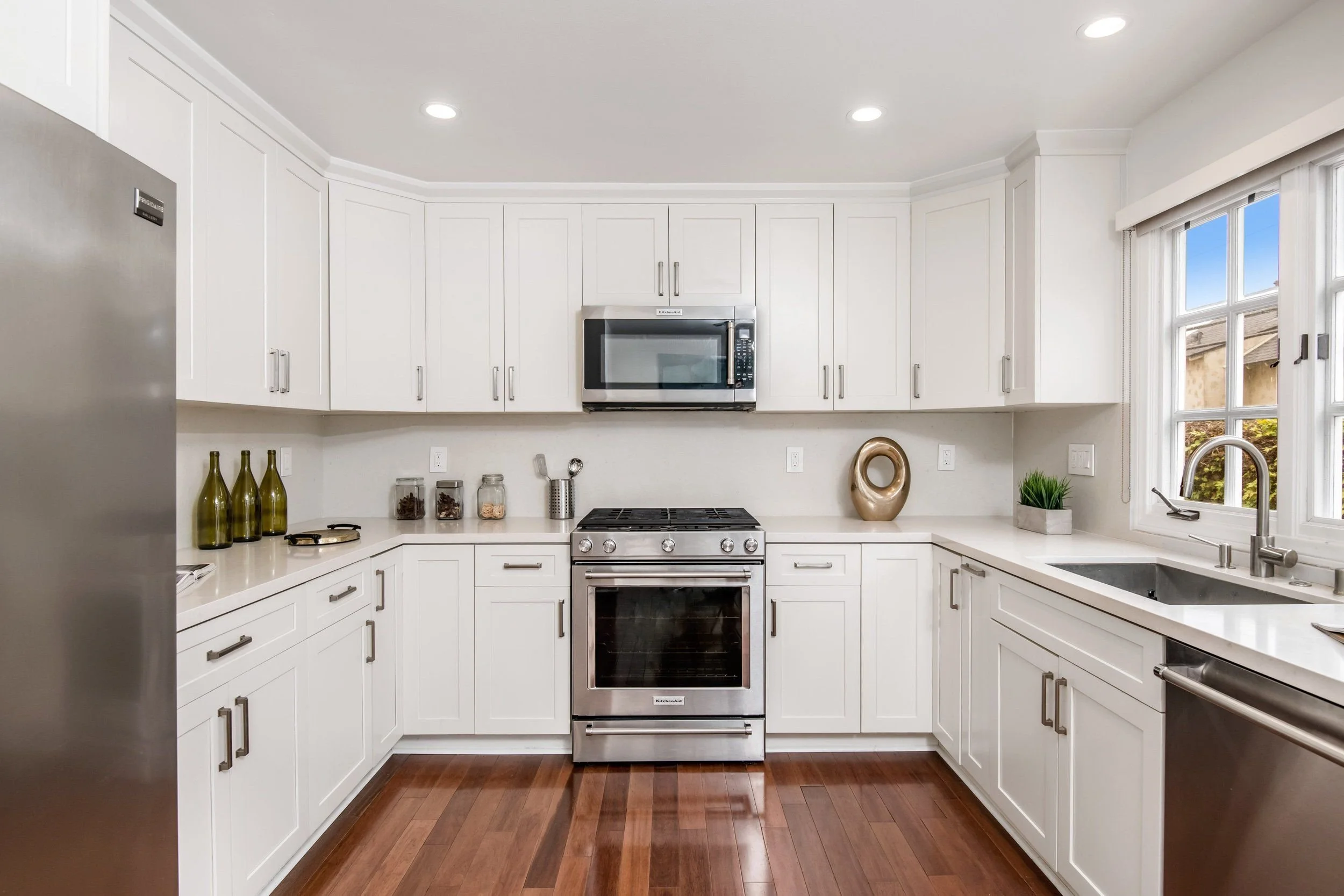 Modern white kitchen with built-in microwave, oven, stainless steel refrigerator, and cabinetry. Countertop decorations include bottles, jars, a decorative sculpture, and potted plant. Large window with vertical blinds overlooks outdoor scene.