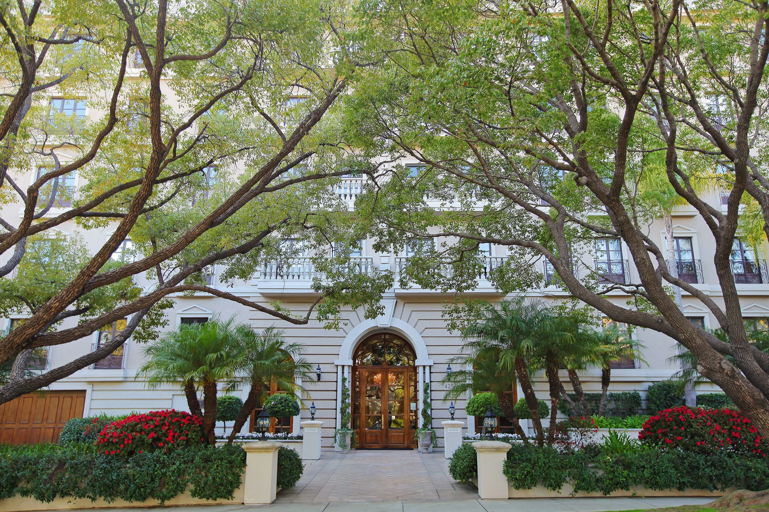 Front view of an elegant residential building with a grand wooden door, surrounded by lush green trees, bushes, and vibrant flowers, creating a welcoming entrance.