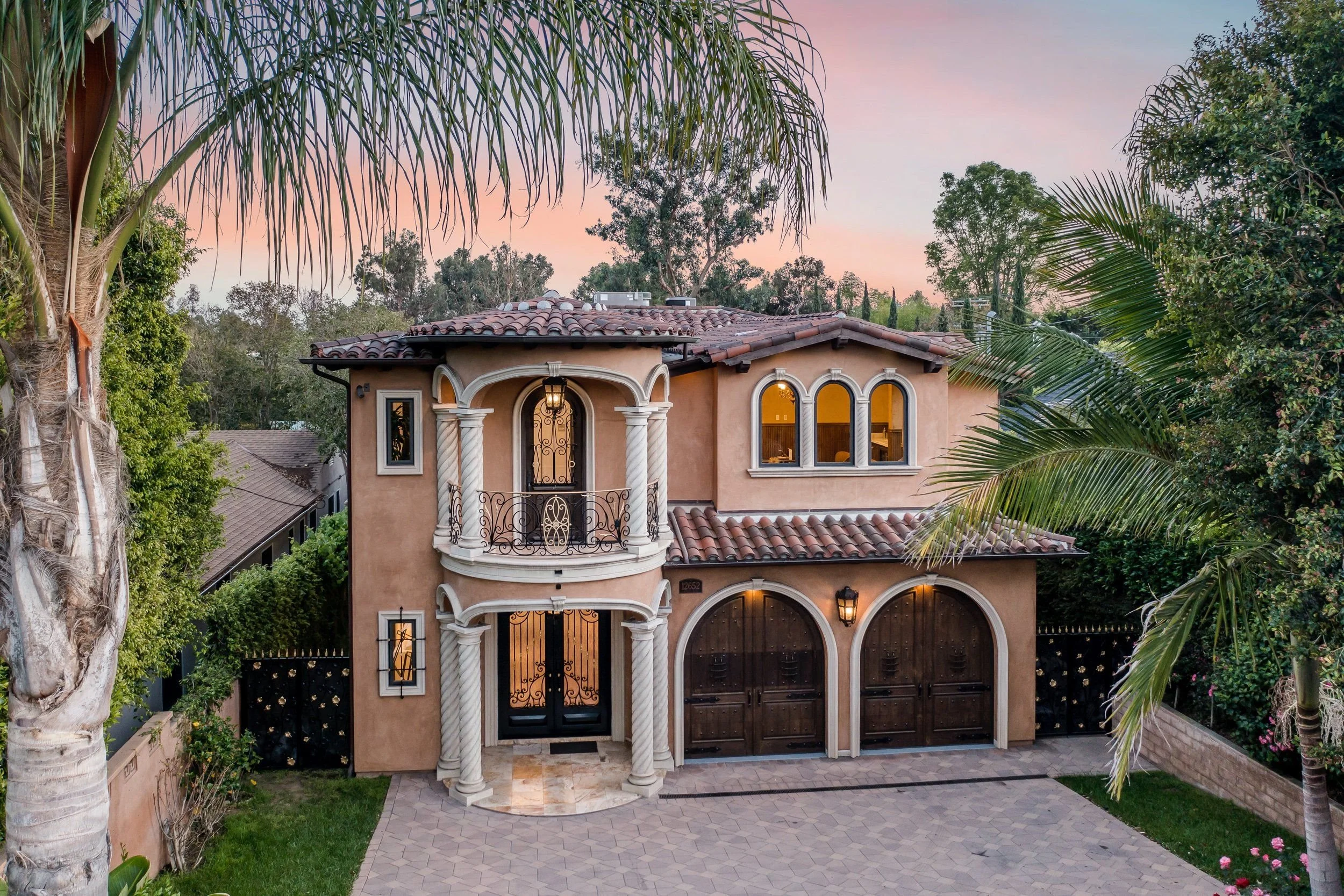 Front view of a two-story house with tropical landscaping and palm trees, sunset sky in the background.