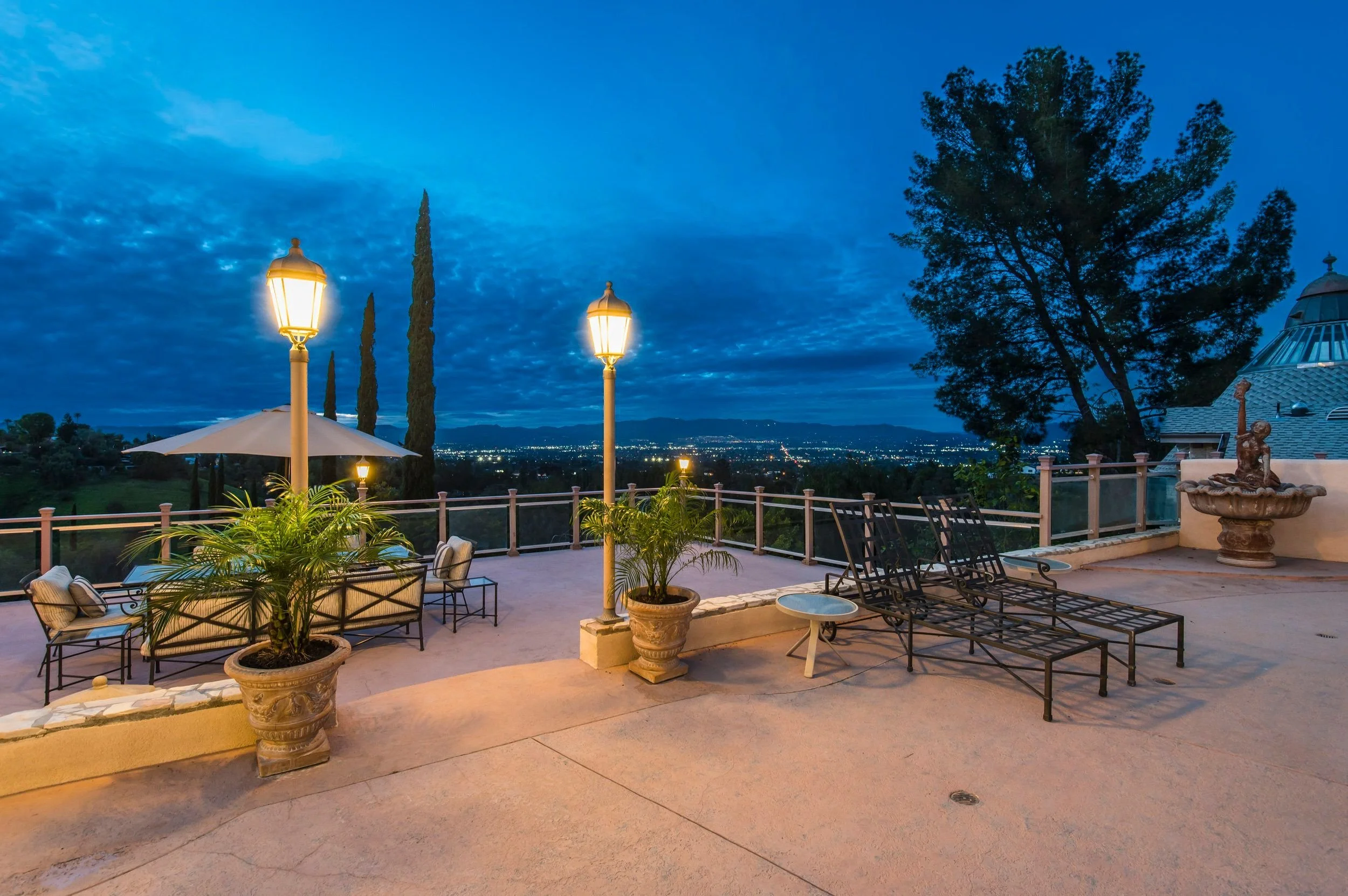 A rooftop terrace at dusk with outdoor furniture, potted plants, an umbrella, and decorative lampposts overlooking a cityscape and distant mountains.