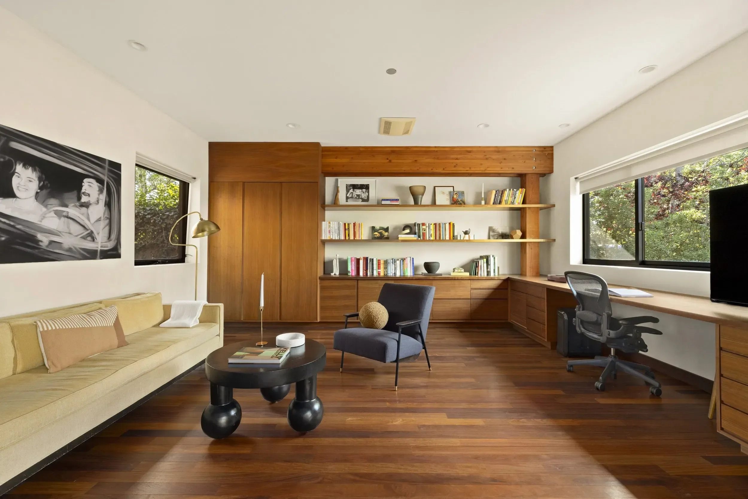 A modern living room with large windows showing trees outside. The room has a beige sofa, a black round coffee table, two chairs, built-in wooden shelves with books and decorative items, and a wooden ceiling beam.