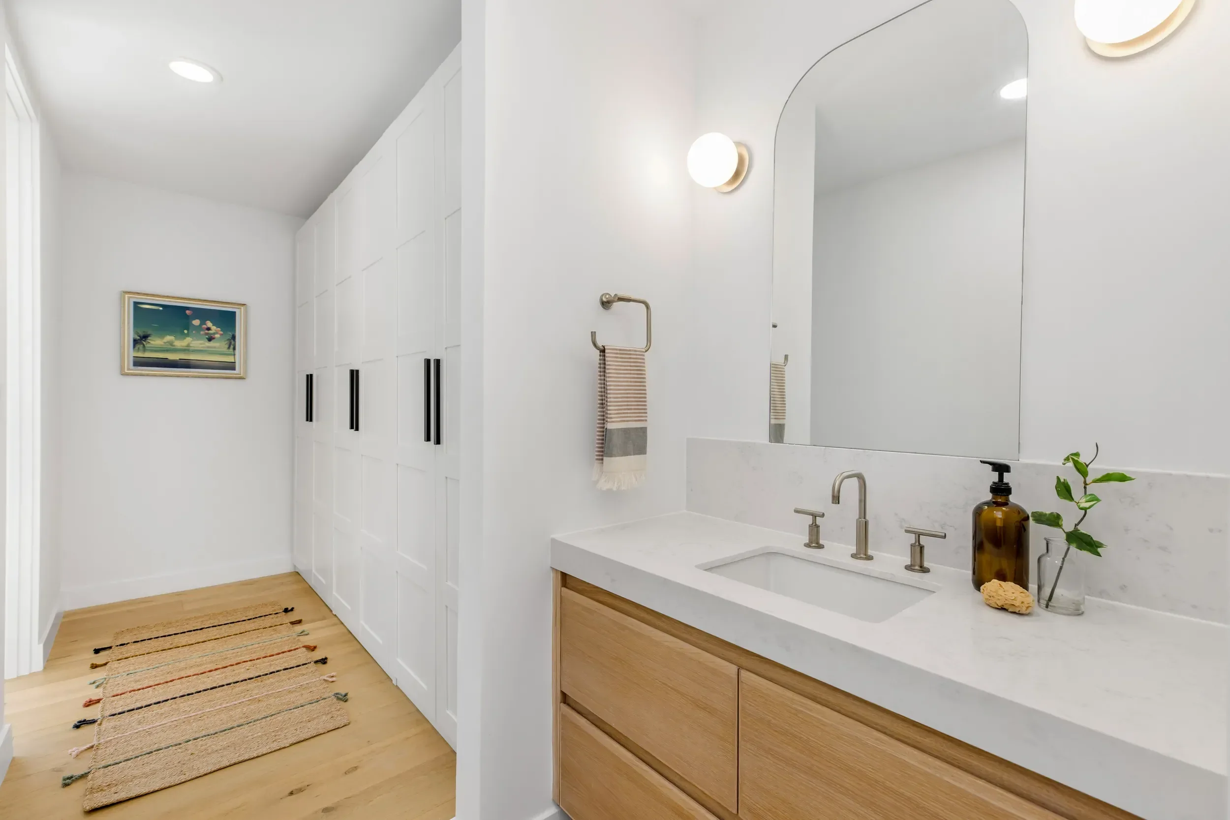 Modern bathroom with white walls, wooden flooring, a large mirror, a white vanity with a black faucet, a wall-mounted light fixture, a towel hanging on a hook, natural decor including a plant and bottles, and a framed artwork with palm trees and a pi