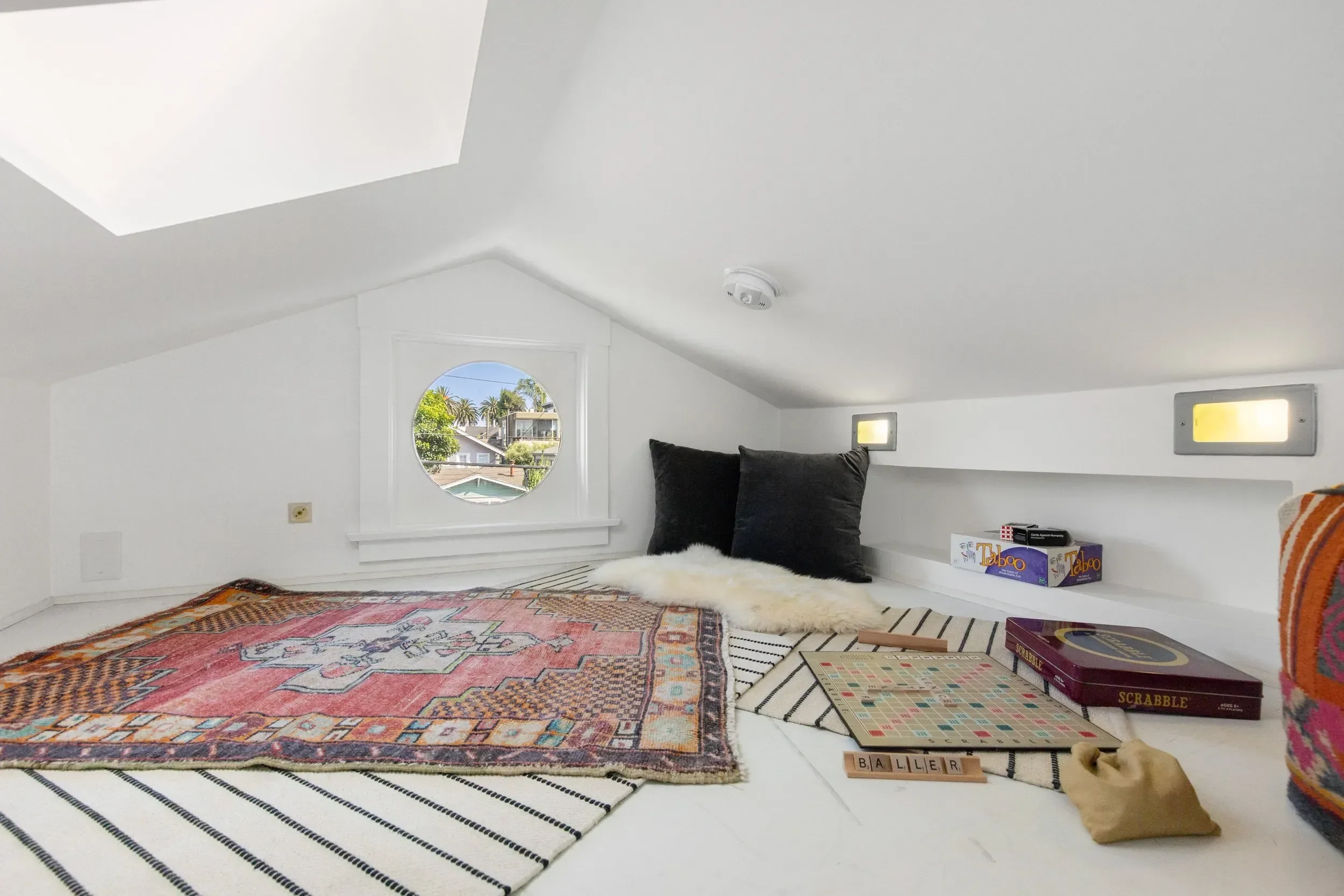 A cozy attic bedroom with white walls and sloped ceiling, featuring a window with a view of palm trees and houses. There is a large colorful area rug on the floor, a fluffy white throw, and black pillows on a built-in bed ledge. Board games including