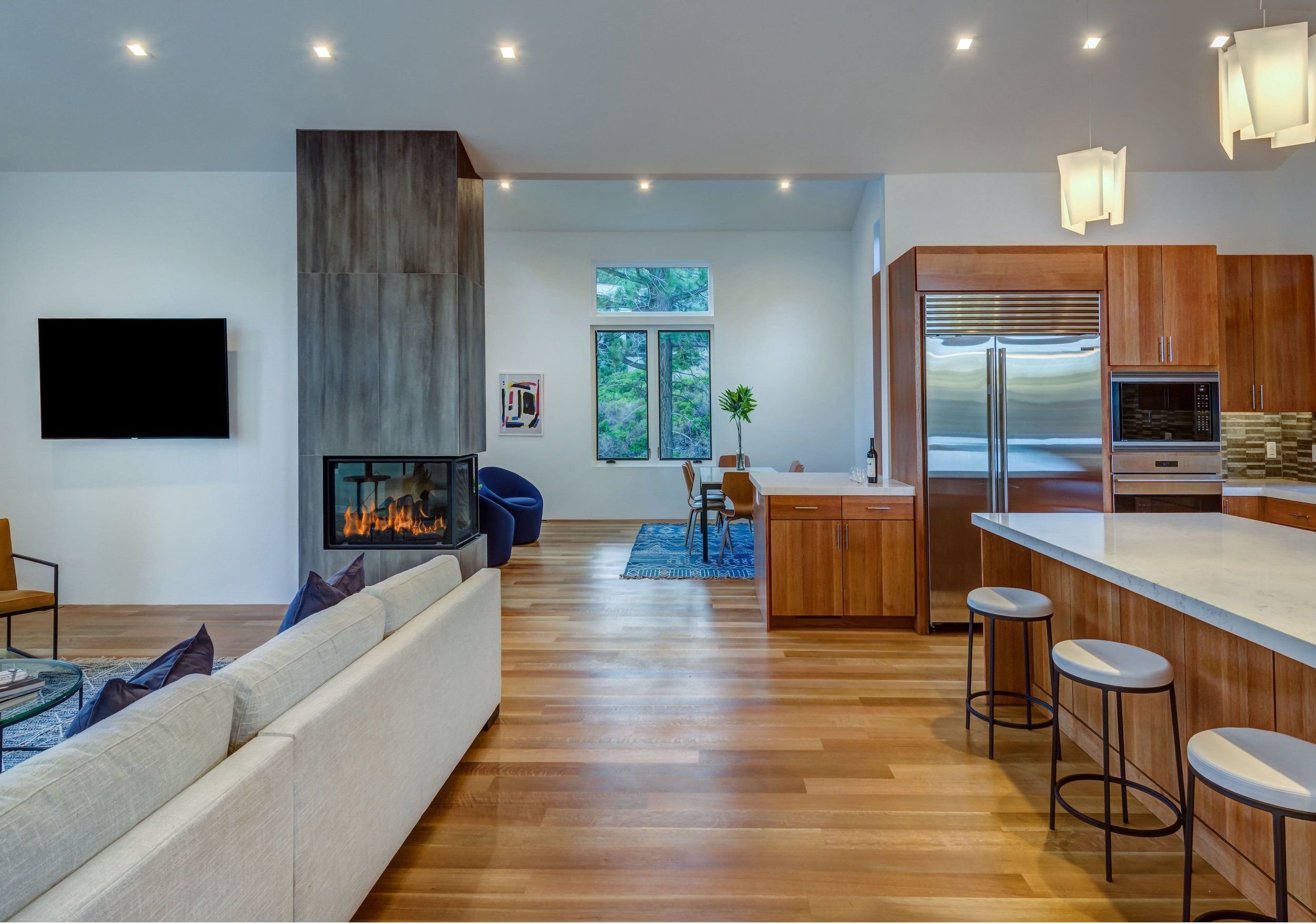 Interior view of a modern open-concept living and kitchen area with a white sofa, a fireplace, a dining table, kitchen cabinets, and appliances, with large windows and wooden flooring.