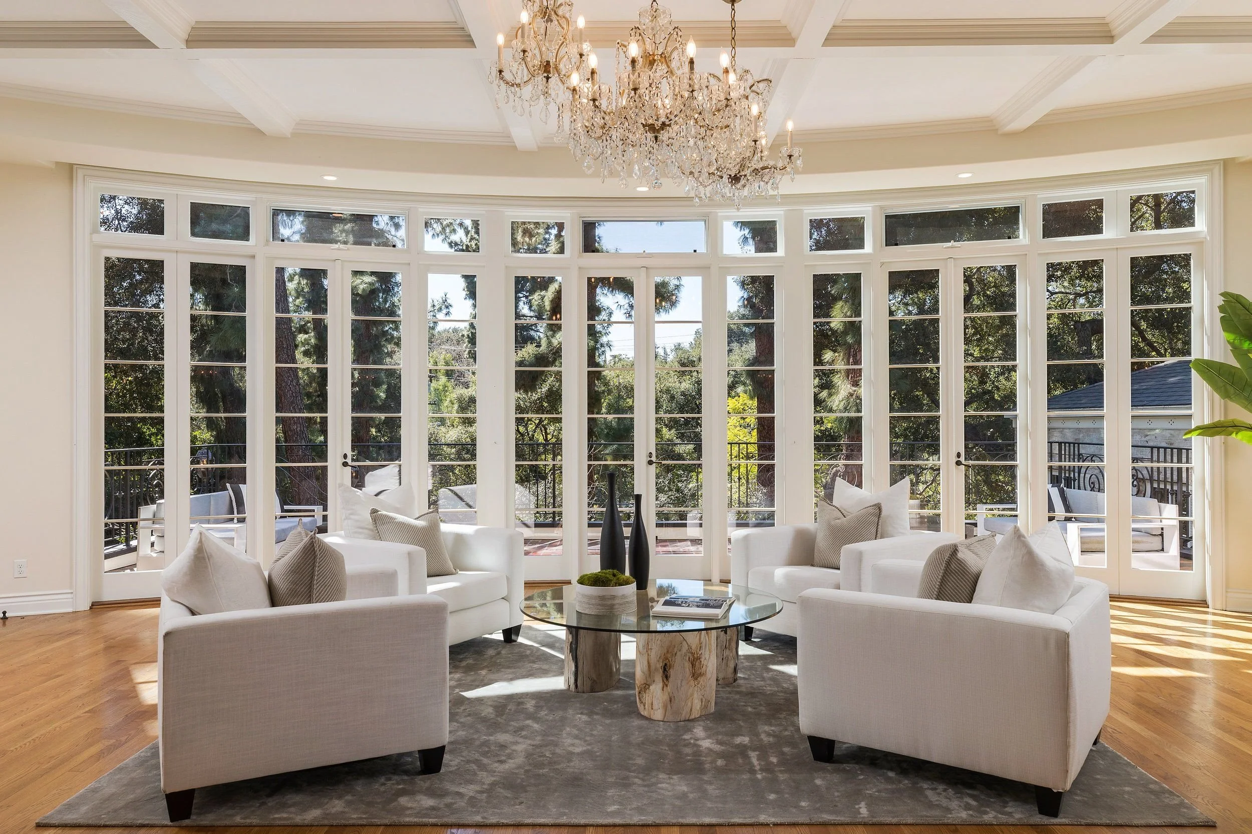 Living room with large circular window, white sofa set, and a glass coffee table on a gray rug. Crystal chandelier hanging from the ceiling and trees visible outside through the windows.