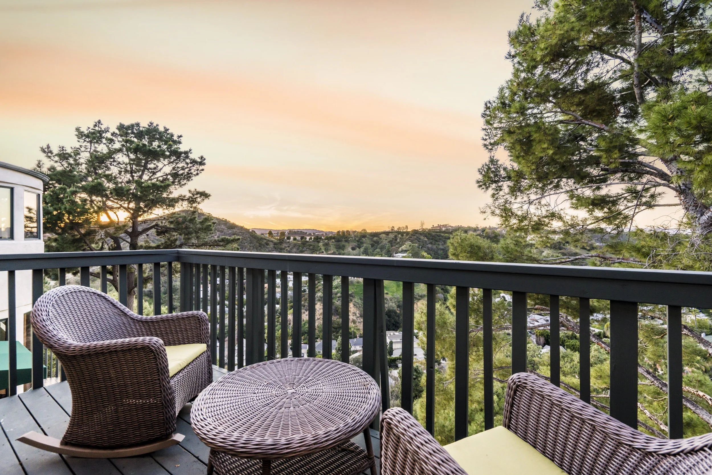 Balcony patio with wicker chairs and table, wooden railing, view of green hills and trees at sunset.