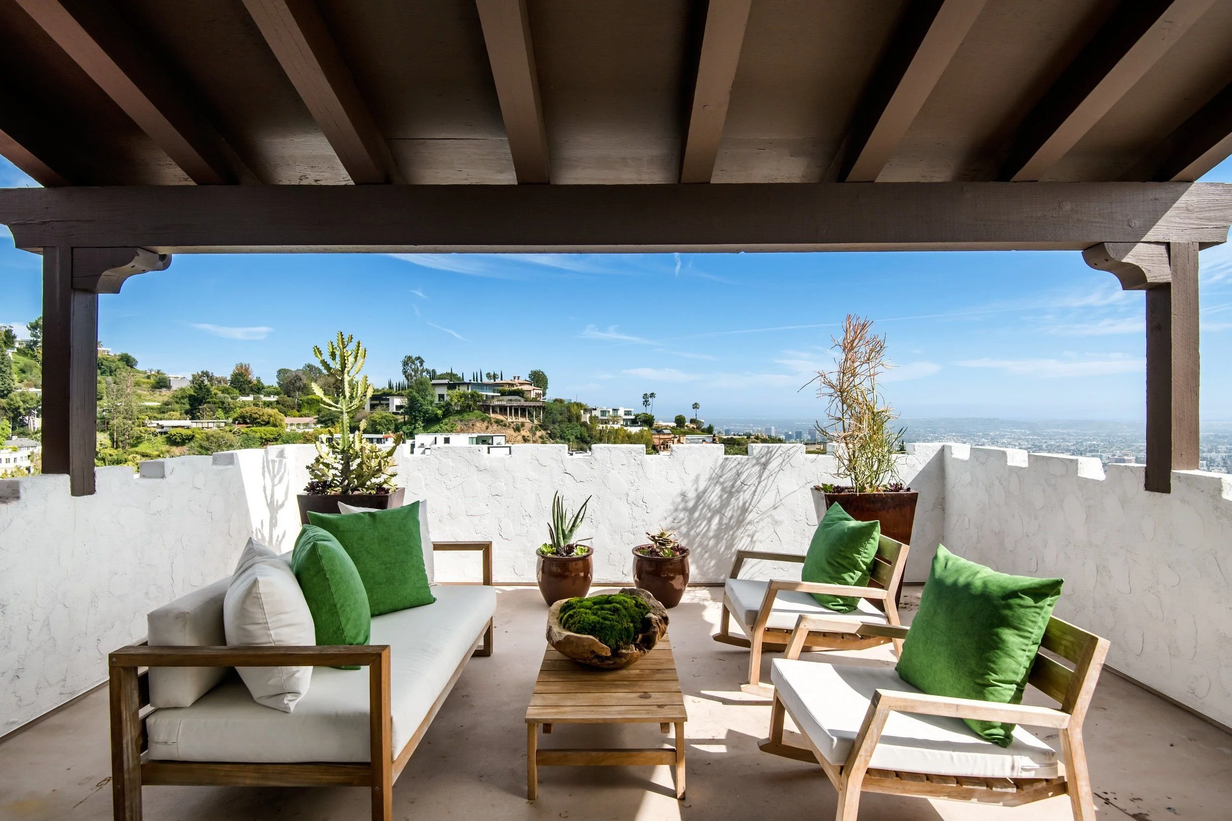 Outdoor patio or balcony with white wall, potted plants, wooden furniture, green cushions, under a wooden pergola, overlooking hilly landscape with houses and blue sky.