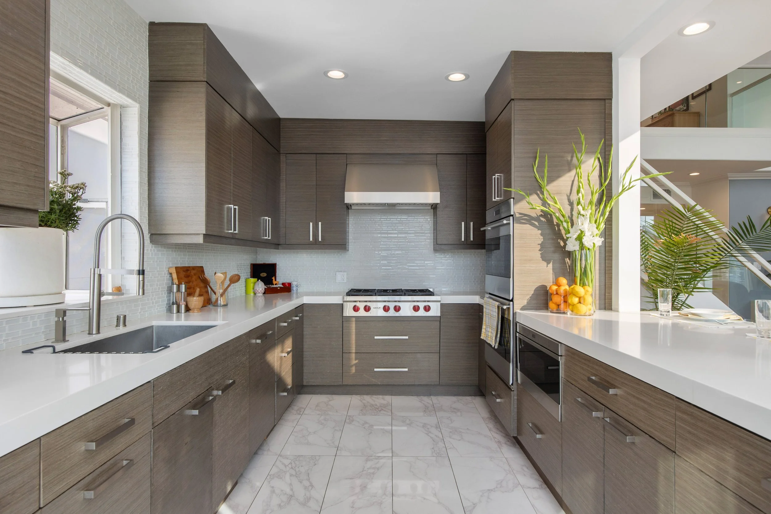 Modern kitchen with wooden cabinetry, white countertops, and stainless steel appliances. There is a window above the sink, a stove in the center, and a large plant with white flowers and oranges on the right side.