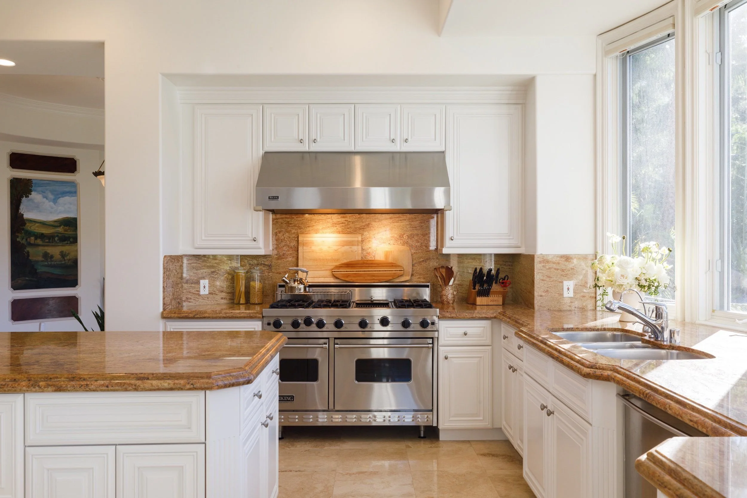 Modern kitchen with white cabinets, granite countertops, stainless steel oven and range hood, large window, and a vase with flowers.