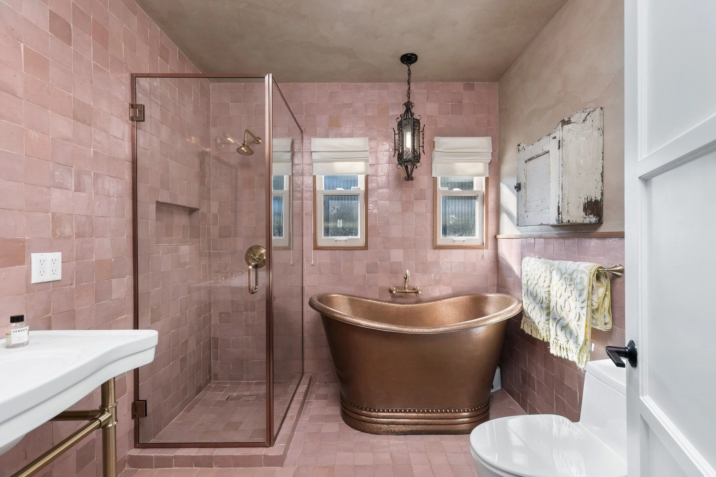 A bathroom with pink tiled walls and floor, a brass tub, a small shower with a glass door, three windows with white shades, a vintage light fixture, a white toilet, and a rustic cabinet on the wall.