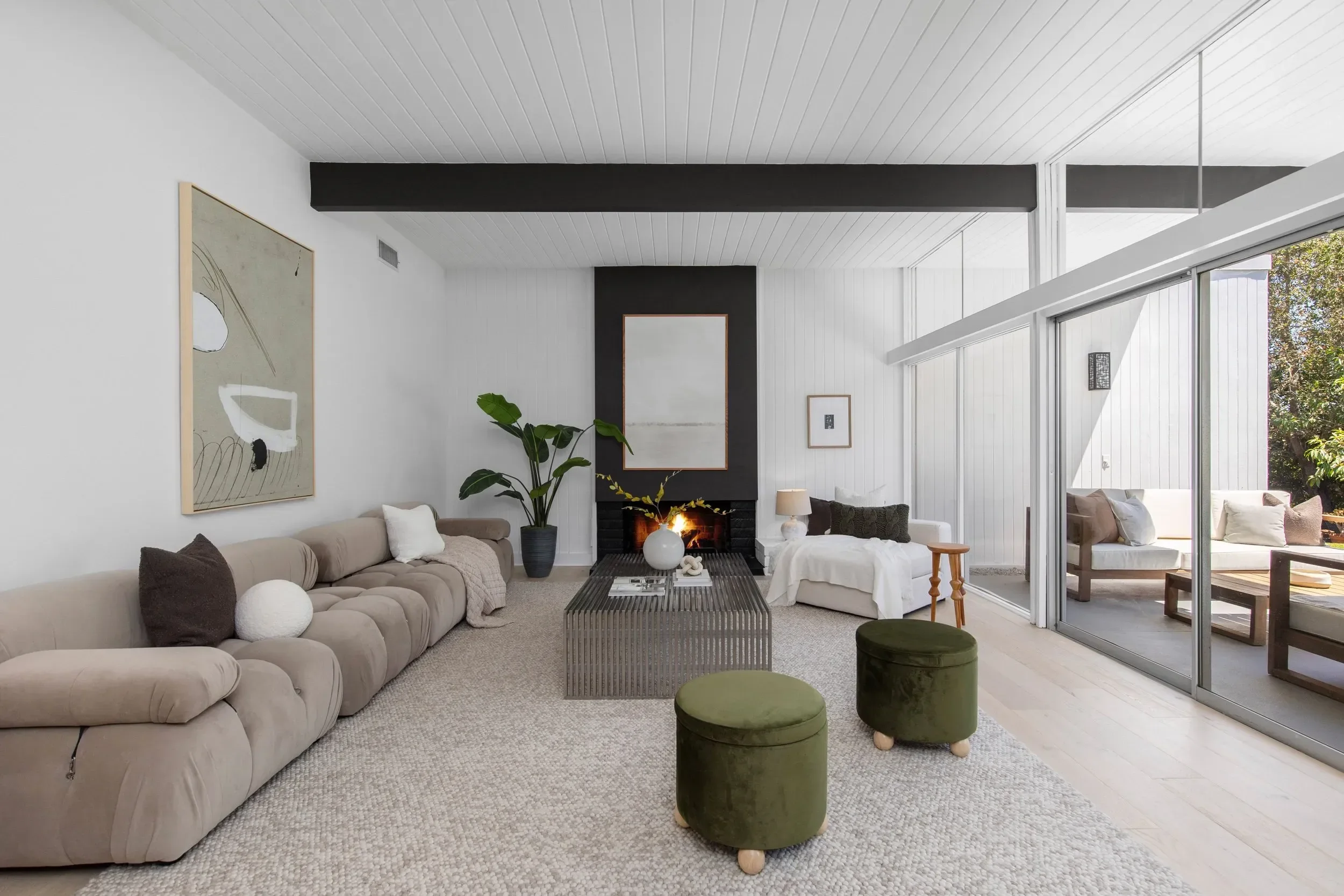 Modern living room with beige sofa, abstract art, green ottomans, fireplace, and large windows showing outdoor patio and greenery.