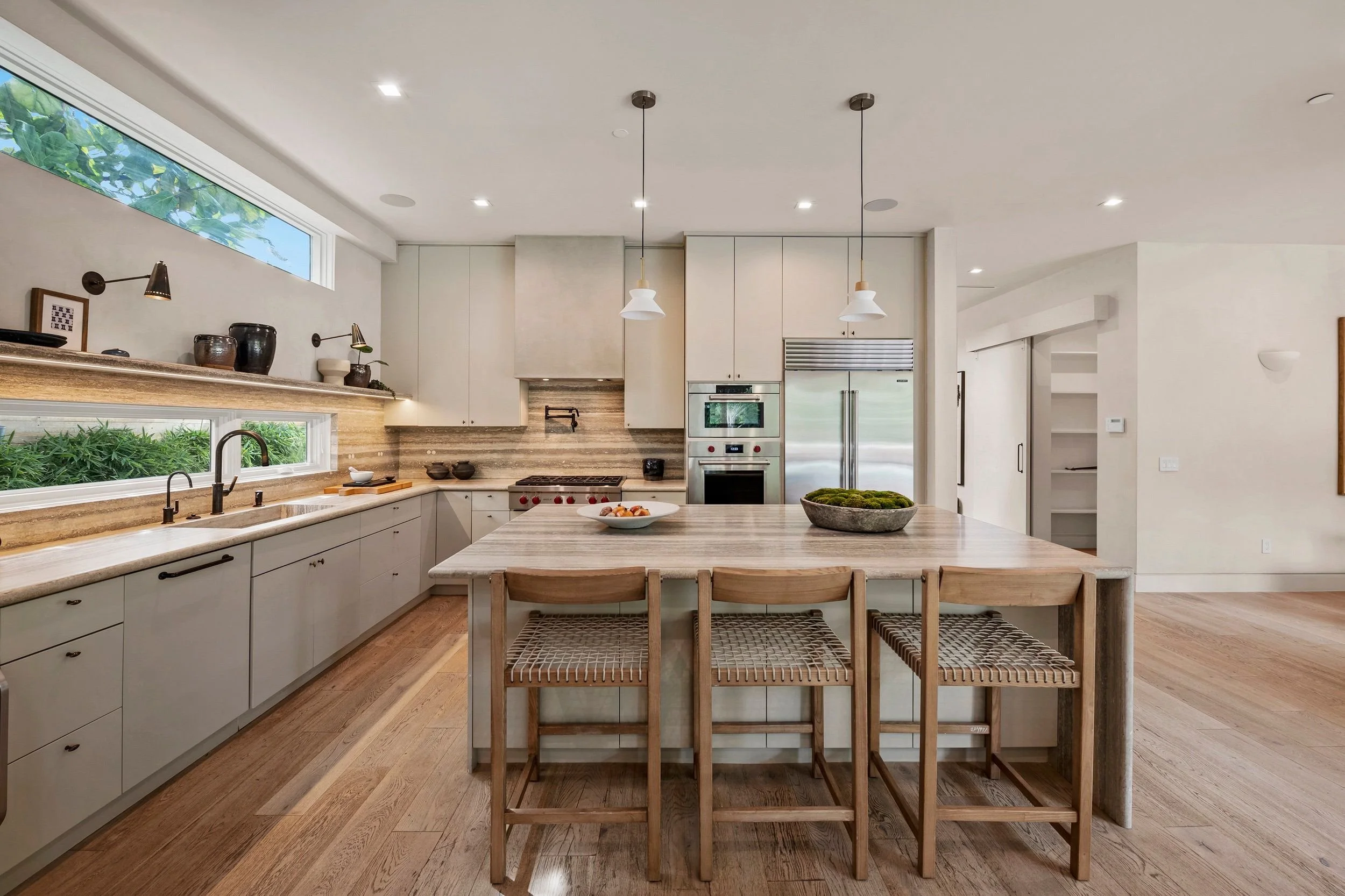 Modern kitchen with white cabinets, wooden countertops, and a kitchen island with four chairs. There are two pendant lights hanging over the island and a window above the sink, with greenery visible outside.